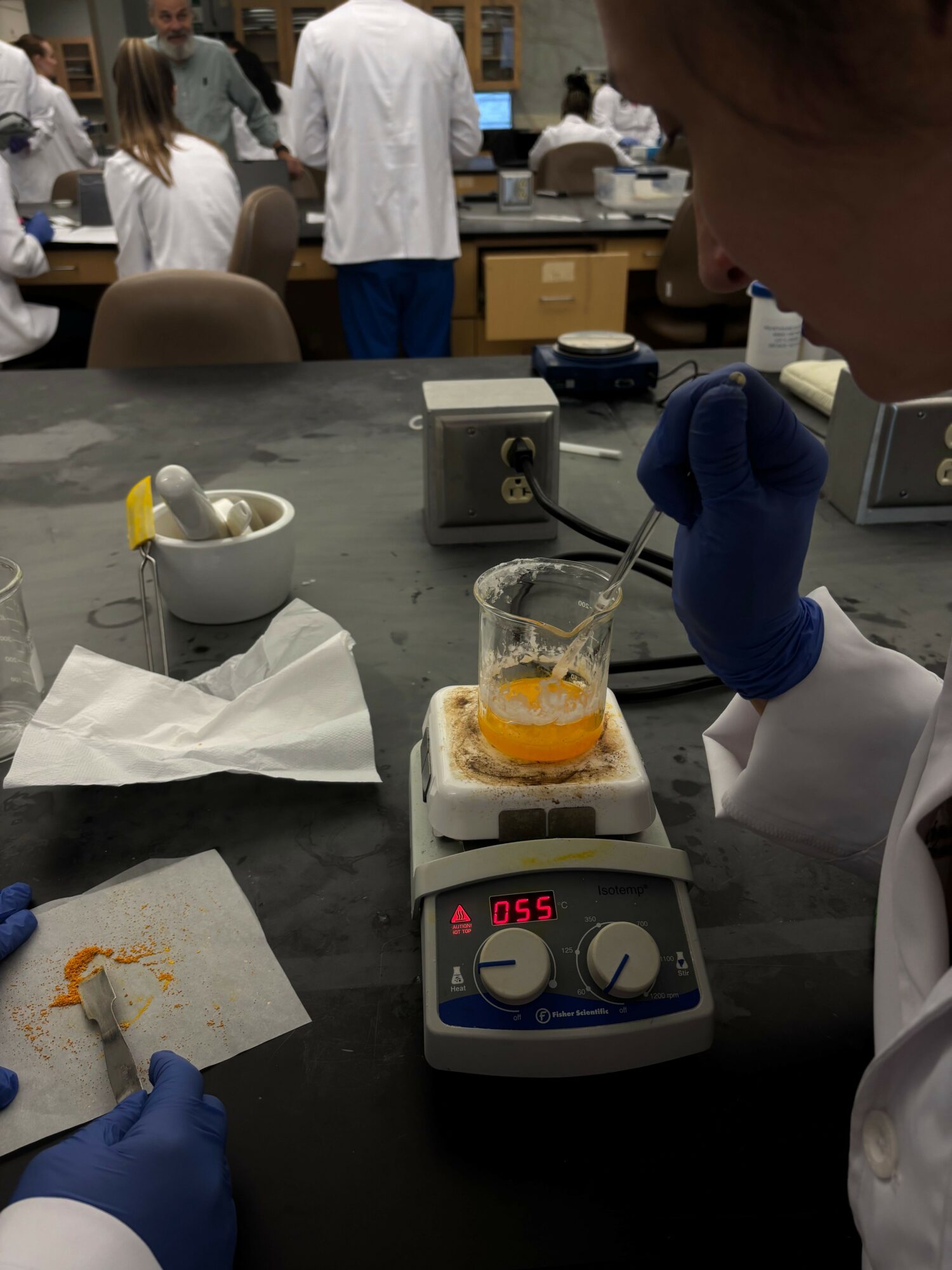 Laboratory scene with a person wearing blue gloves working with a beaker and equipment on a black countertop.