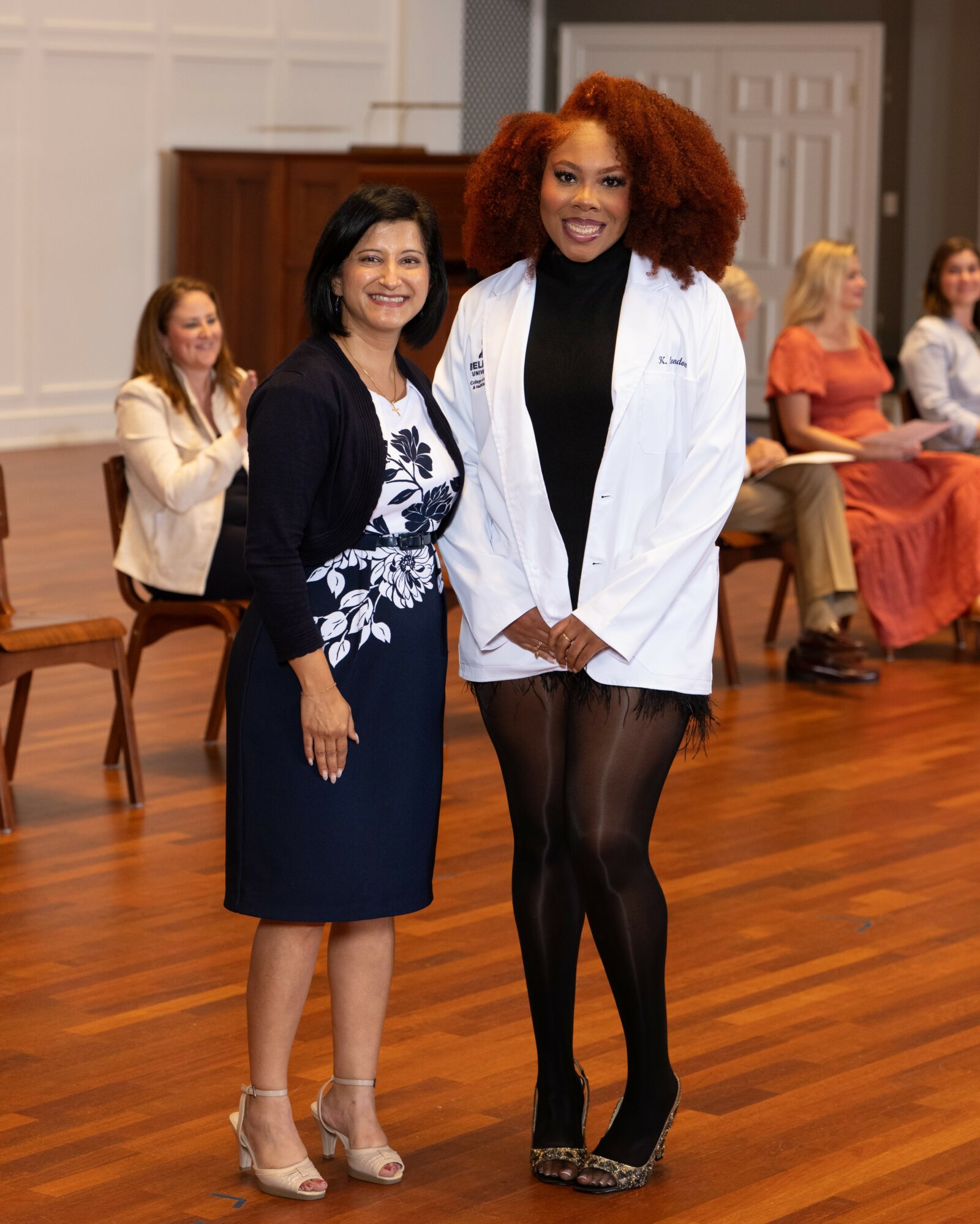 Two women standing together in a room with wooden floors and chairs, smiling at the camera, one in a white coat and the other in a dark dress.