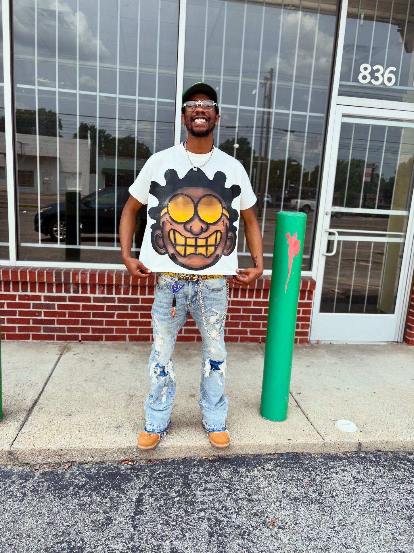 Person standing outside in front of a glass building, wearing a graphic t-shirt, jeans, and sunglasses, smiling.