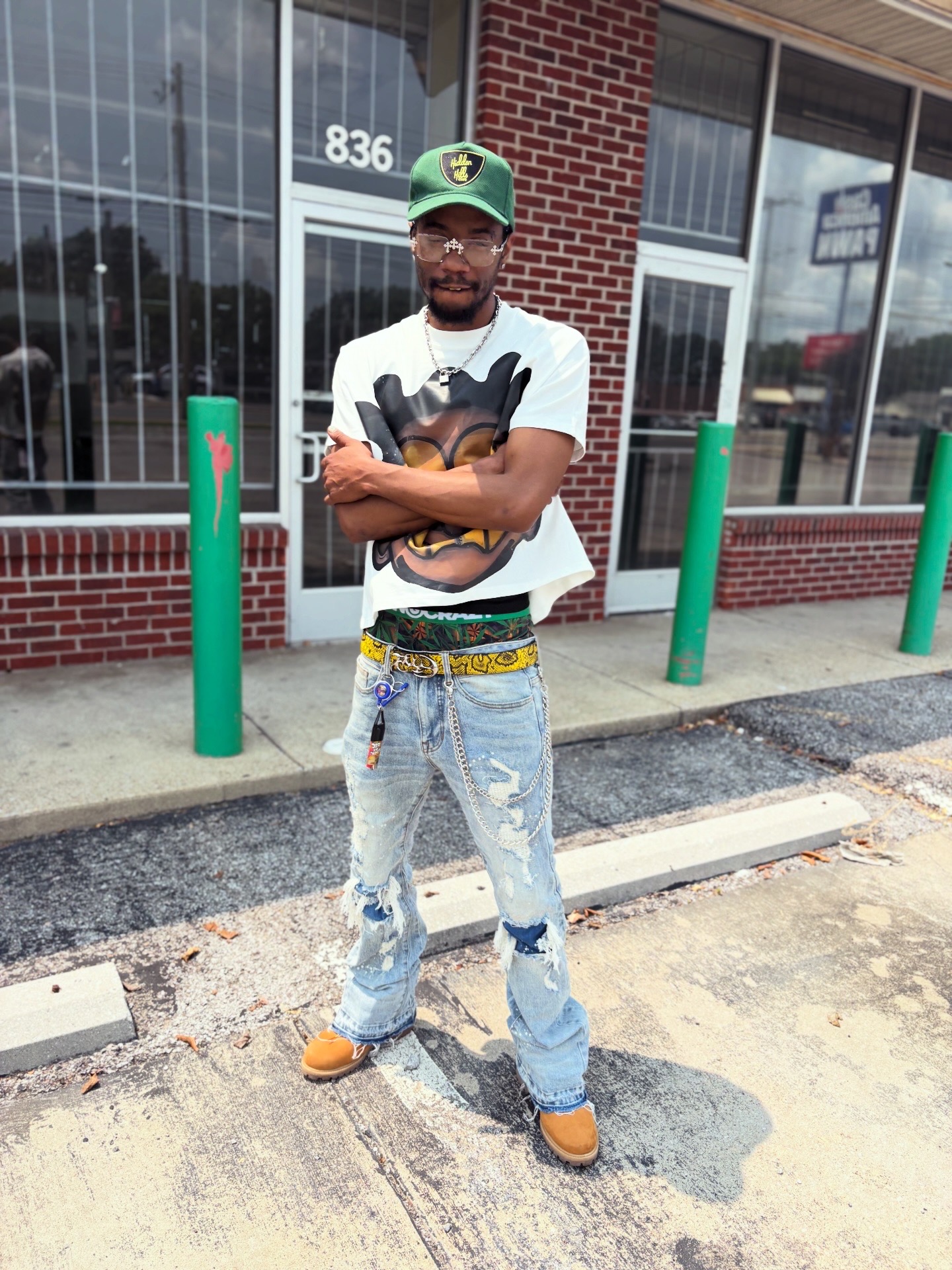 Young man standing outside in front of a brick building with large windows, crossing arms, wearing a green cap, white t-shirt, and ripped jeans.