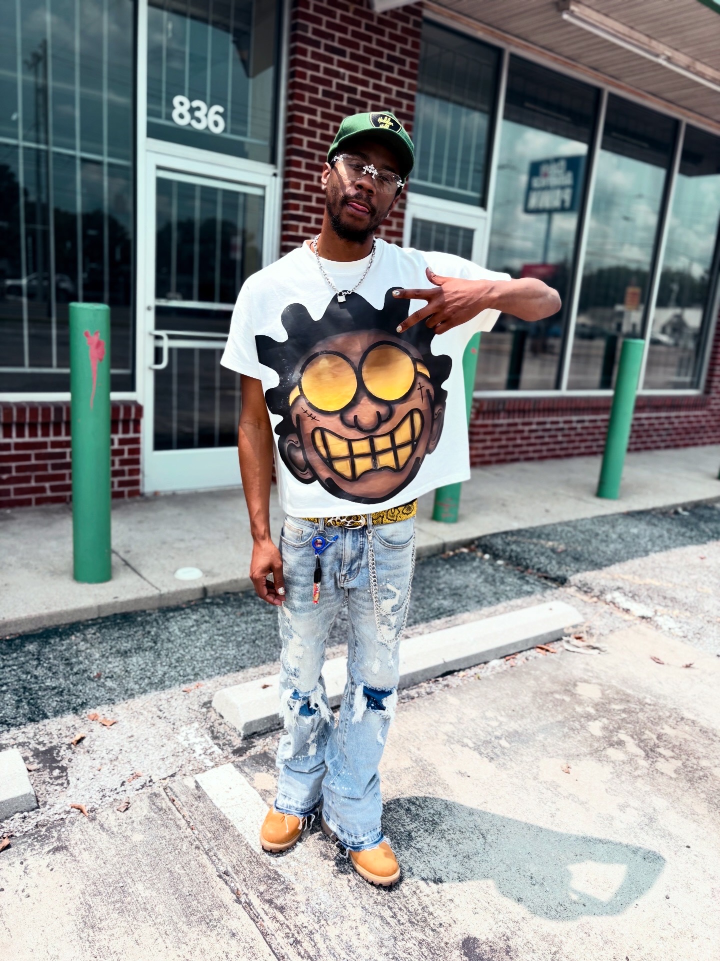 Young man standing outside a building with glass windows, wearing a graphic t-shirt, ripped jeans, and a cap, holding a phone.