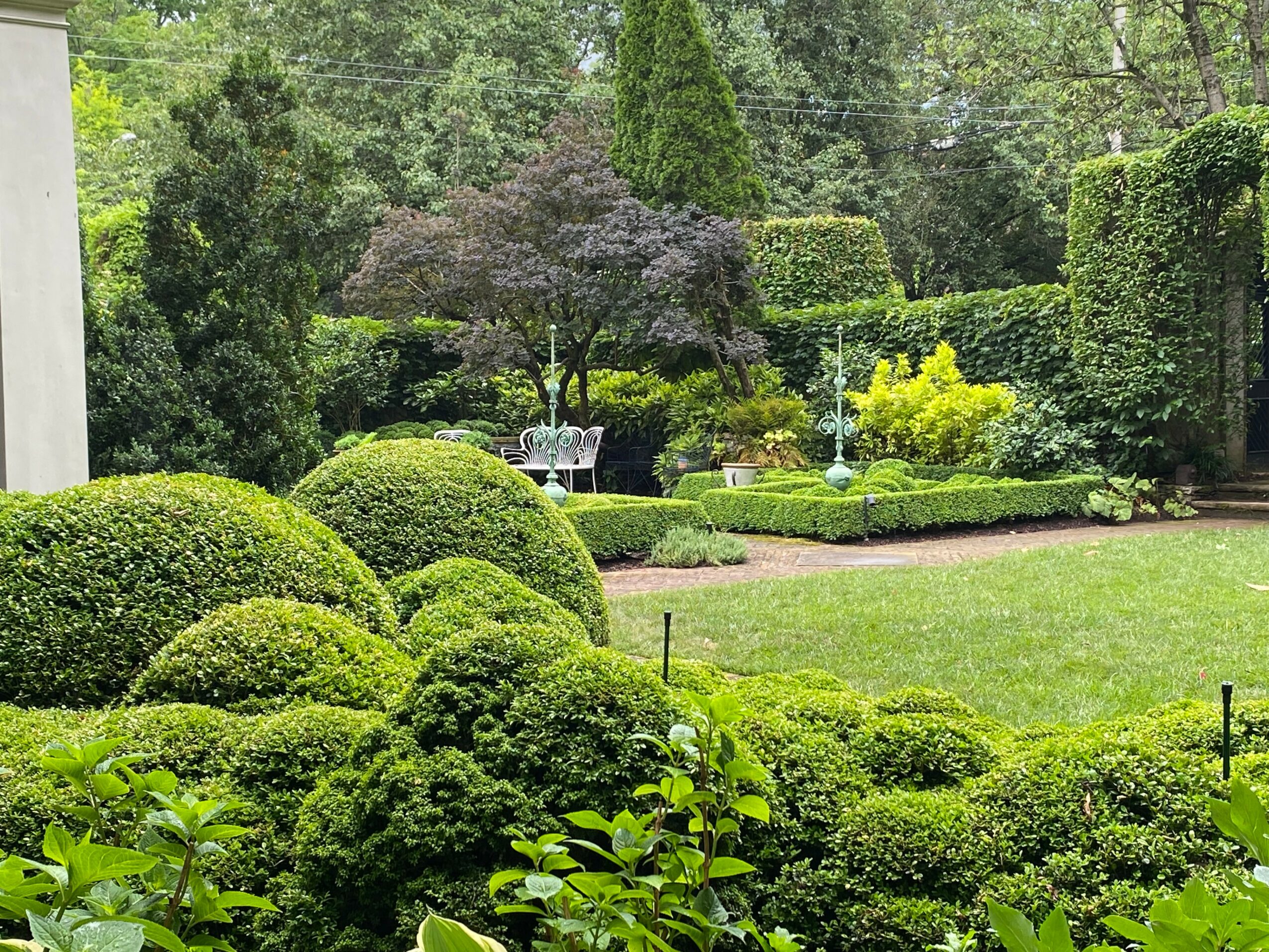 Lush garden with trimmed bushes, trees, and a lawn, featuring a bench and pathway, surrounded by greenery.
