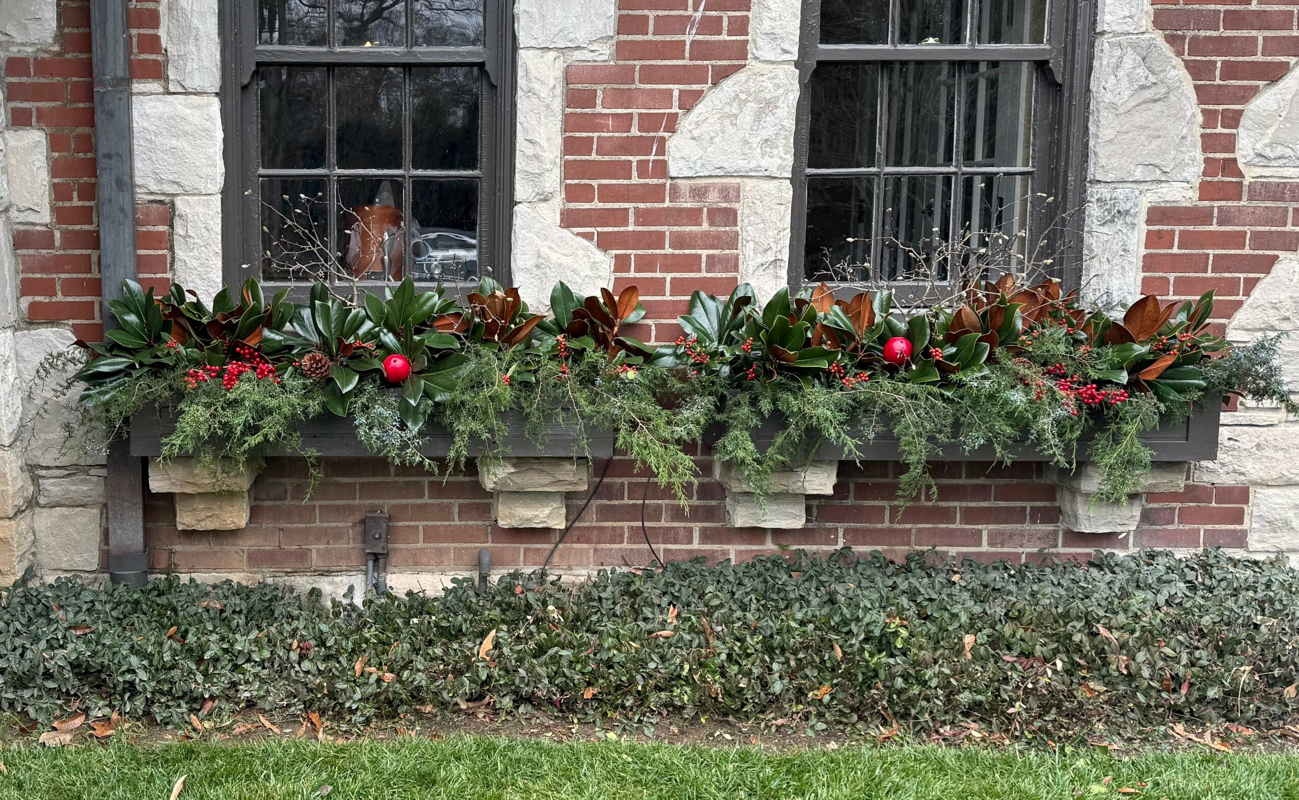 Decorative holiday greenery with red ornaments and pinecones on a brick wall window ledge, with a hedge below.