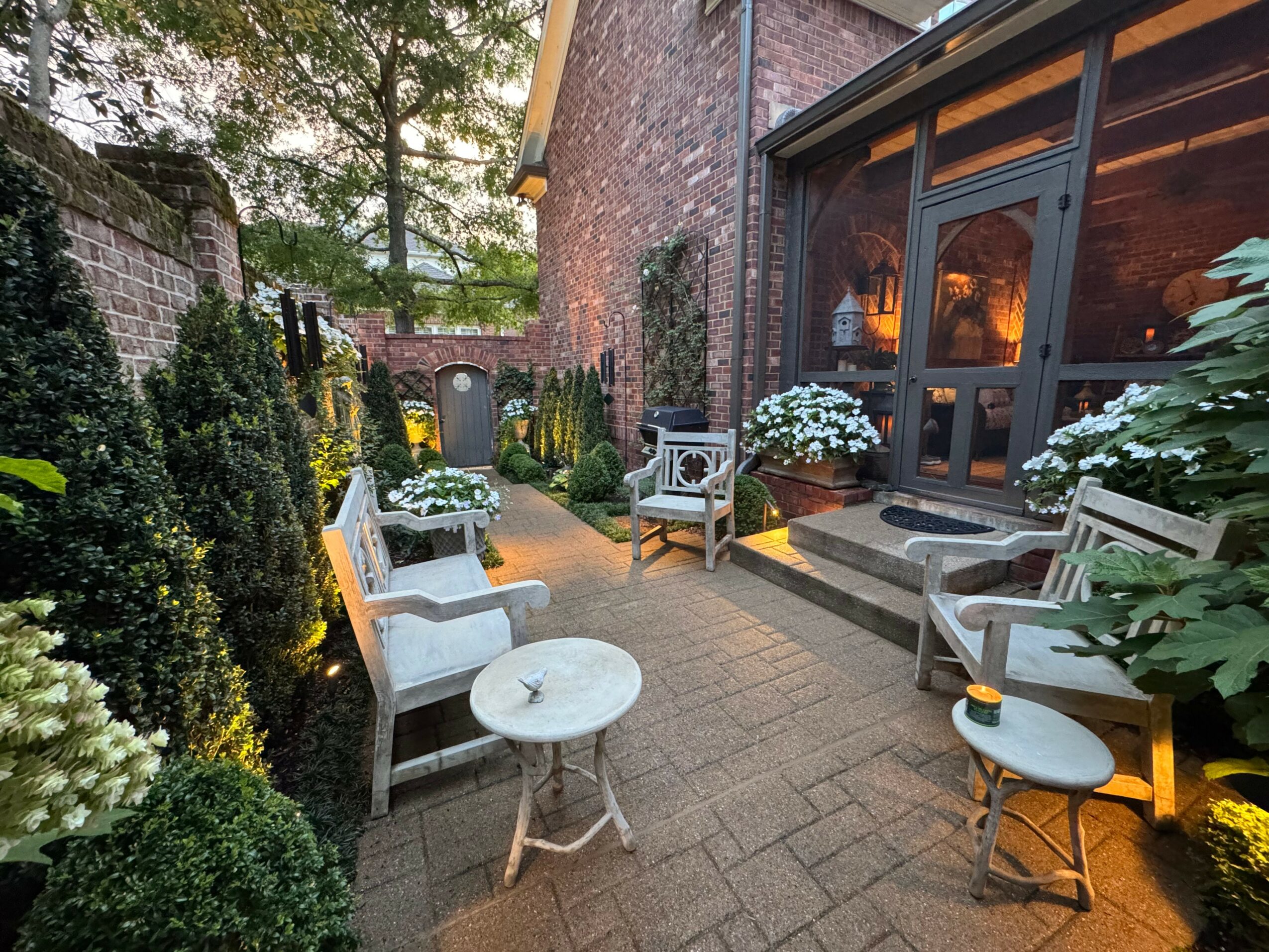 Outdoor patio with chairs, tables, and potted plants near a brick building, illuminated by warm lighting.