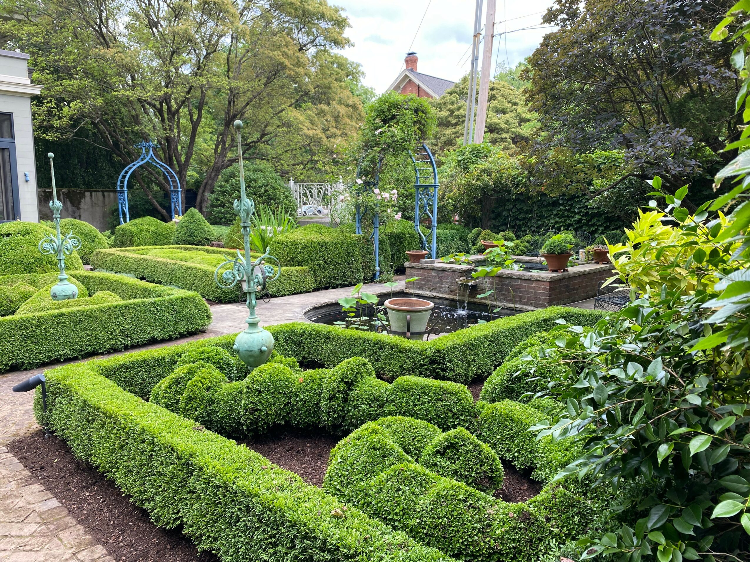 A lush garden with neatly trimmed hedges, decorative blue garden ornaments, and a small fountain area, surrounded by trees and greenery.