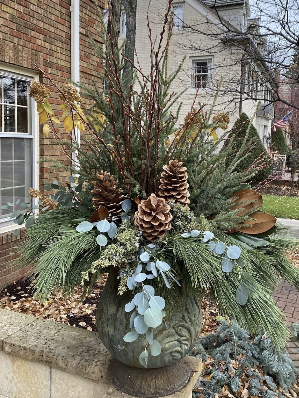 Large floral arrangement with pinecones, eucalyptus, and evergreen branches in a decorative vase outdoors.
