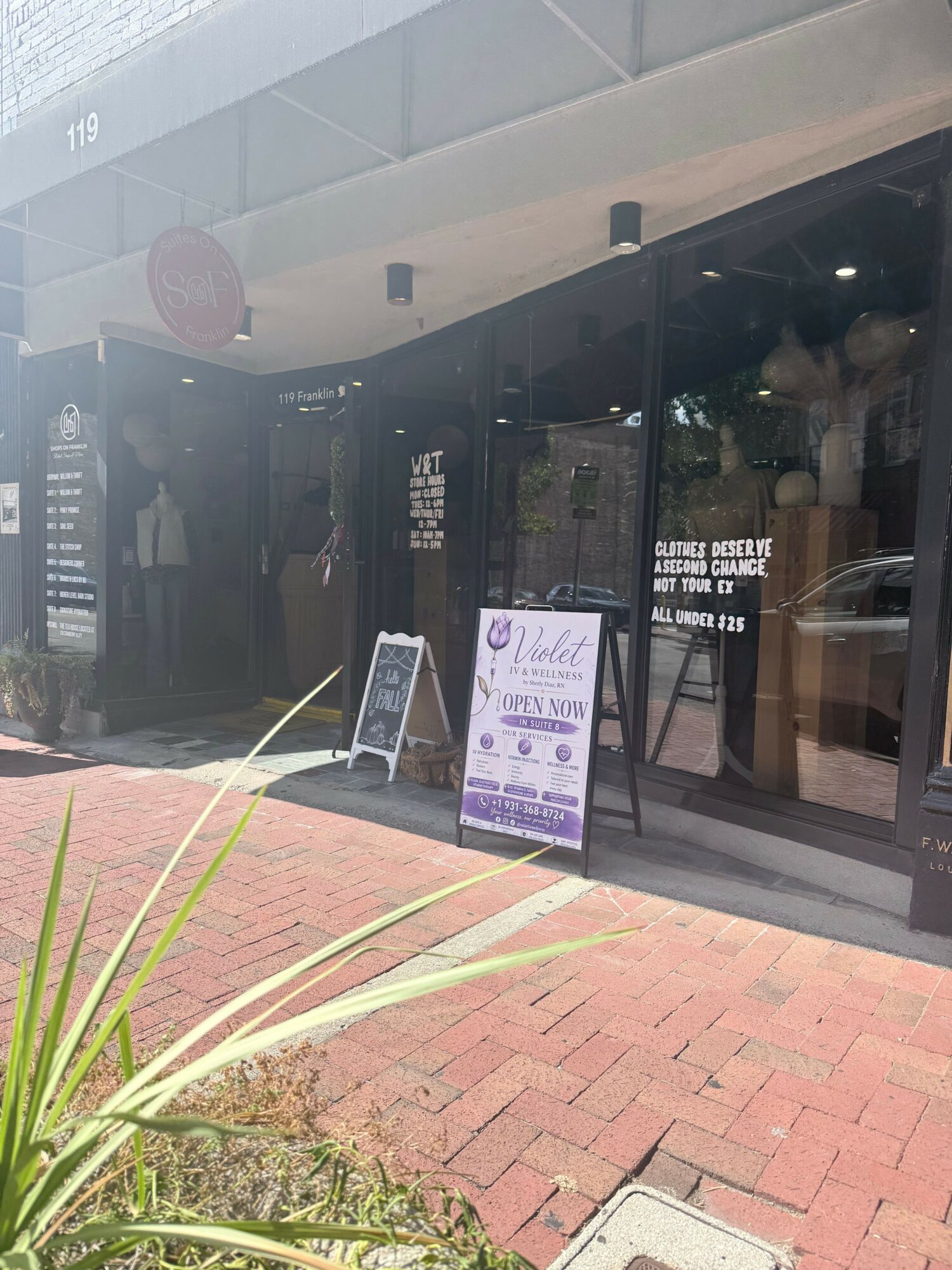 Store entrance with glass door, sign, and potted plant outside on brick sidewalk.