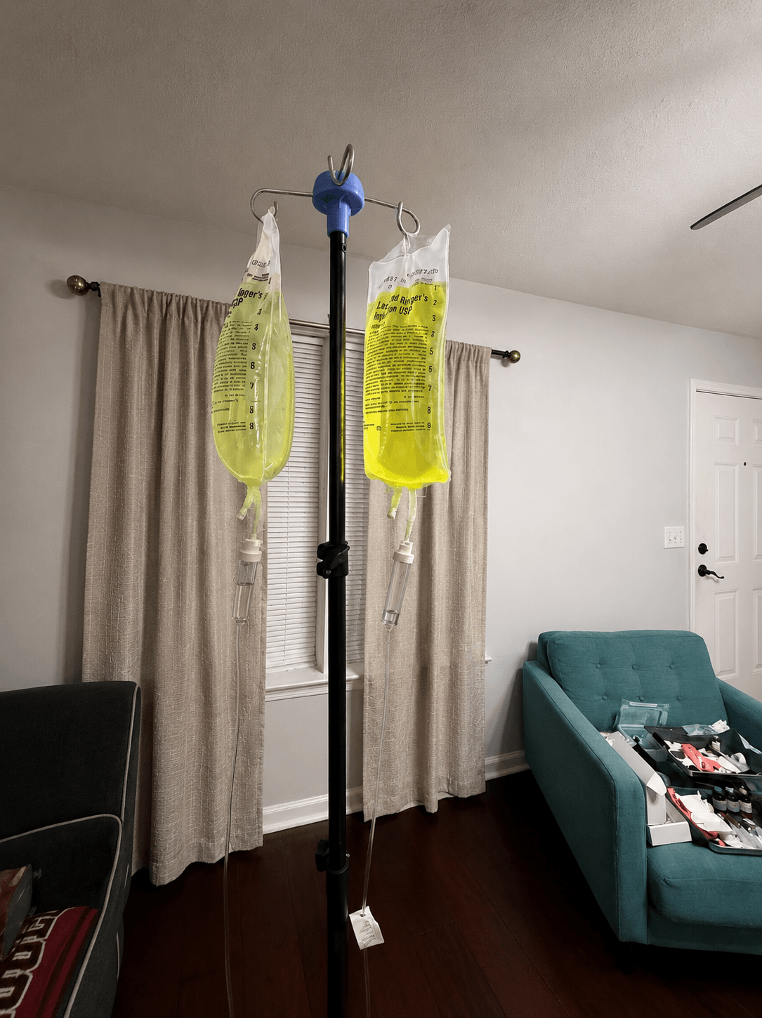 Medical IV pole with two yellow infusion bags hanging in a living room with curtains and furniture