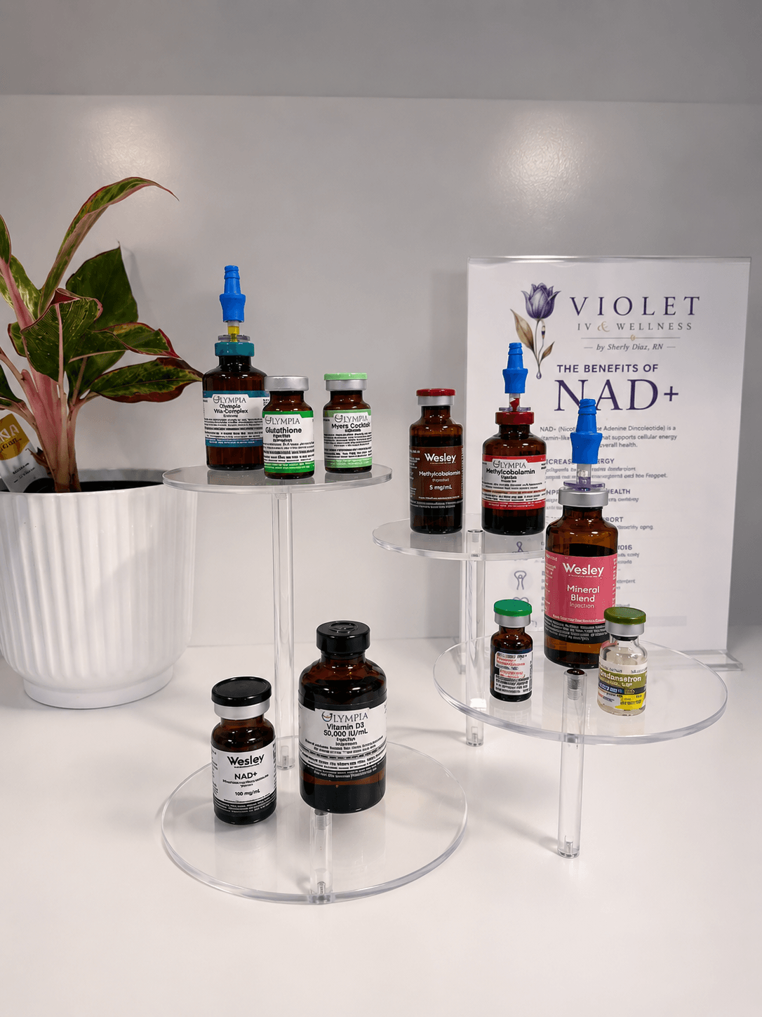 Vials and bottles of medication or supplements displayed on clear circular stands with a potted plant and a sign in the background.