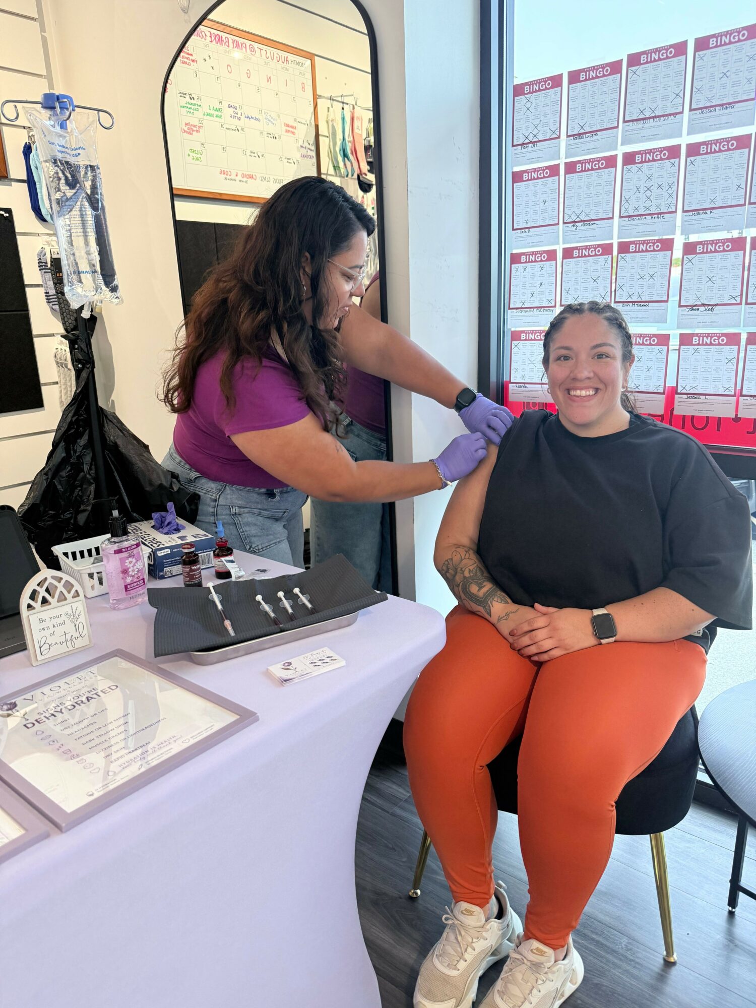 A woman in a purple shirt and gloves is administering a vaccine to a seated woman in black shirt and orange pants, indoors.