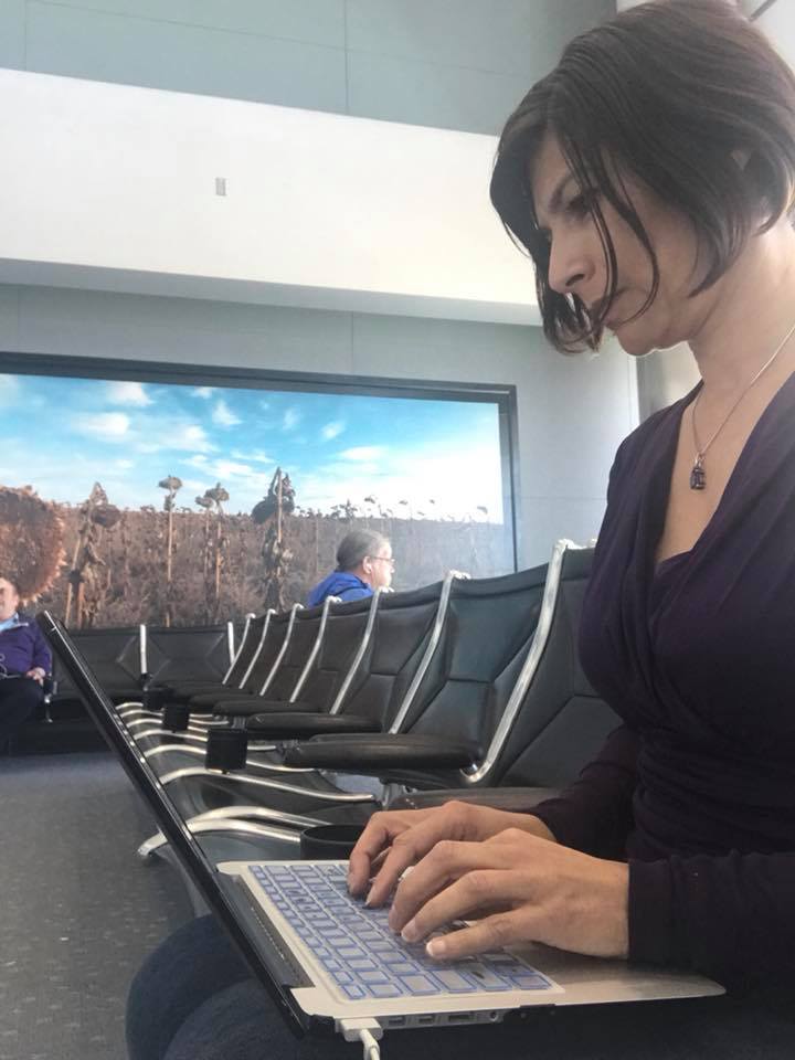 Woman with short dark hair working on a laptop in an airport terminal, with rows of chairs and a large window showing an outdoor scene.