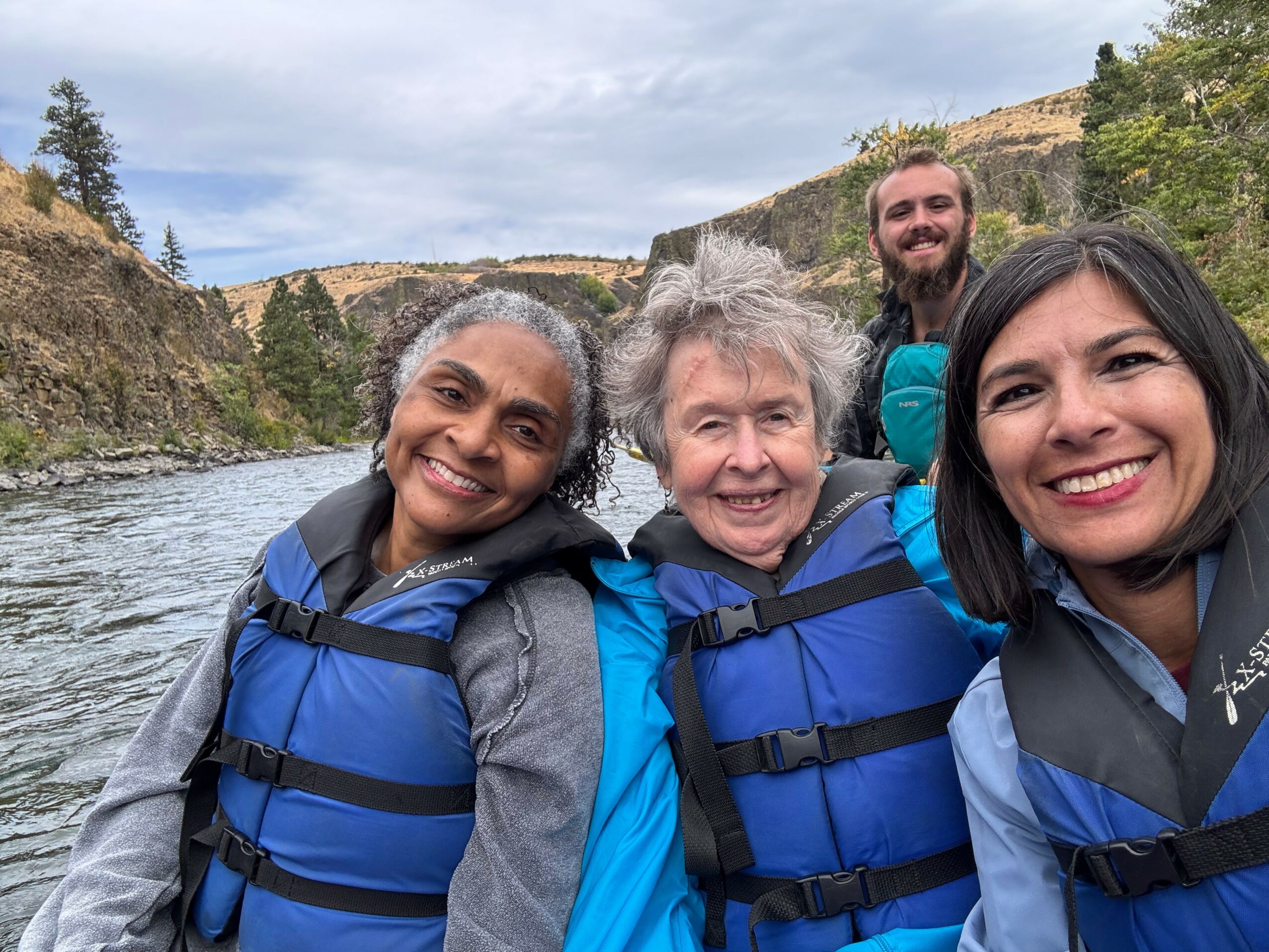 Four people wearing life jackets outdoors near a river with cliffs and trees in the background, smiling at the camera.