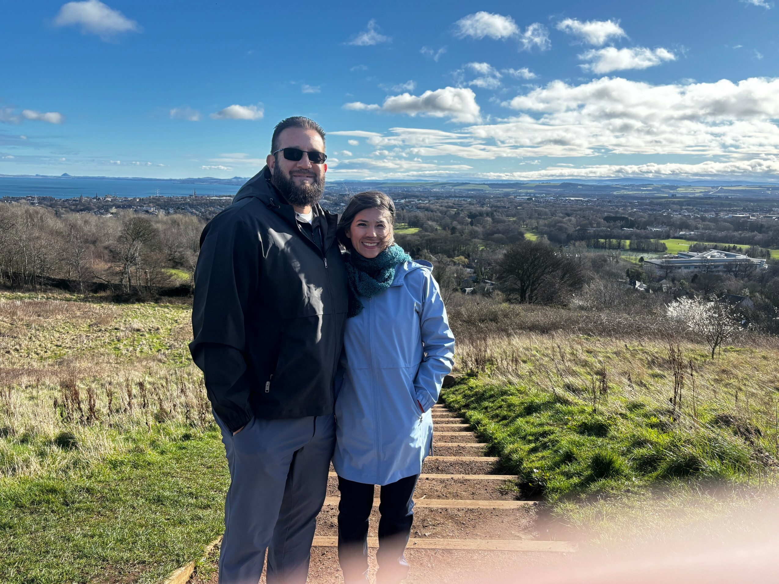 A man and woman stand outdoors on a sunny day with a landscape of fields and trees behind them.