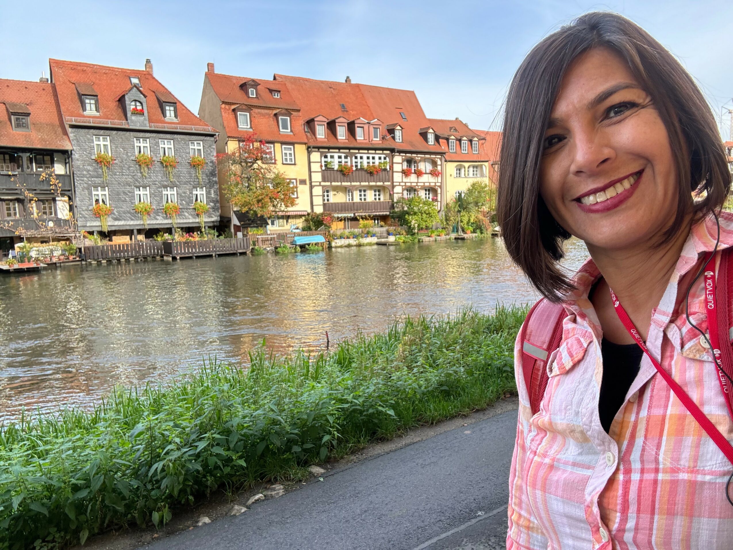 Smiling woman with shoulder-length dark hair wearing a pink and white checkered shirt, standing near a river with colorful buildings in the background.