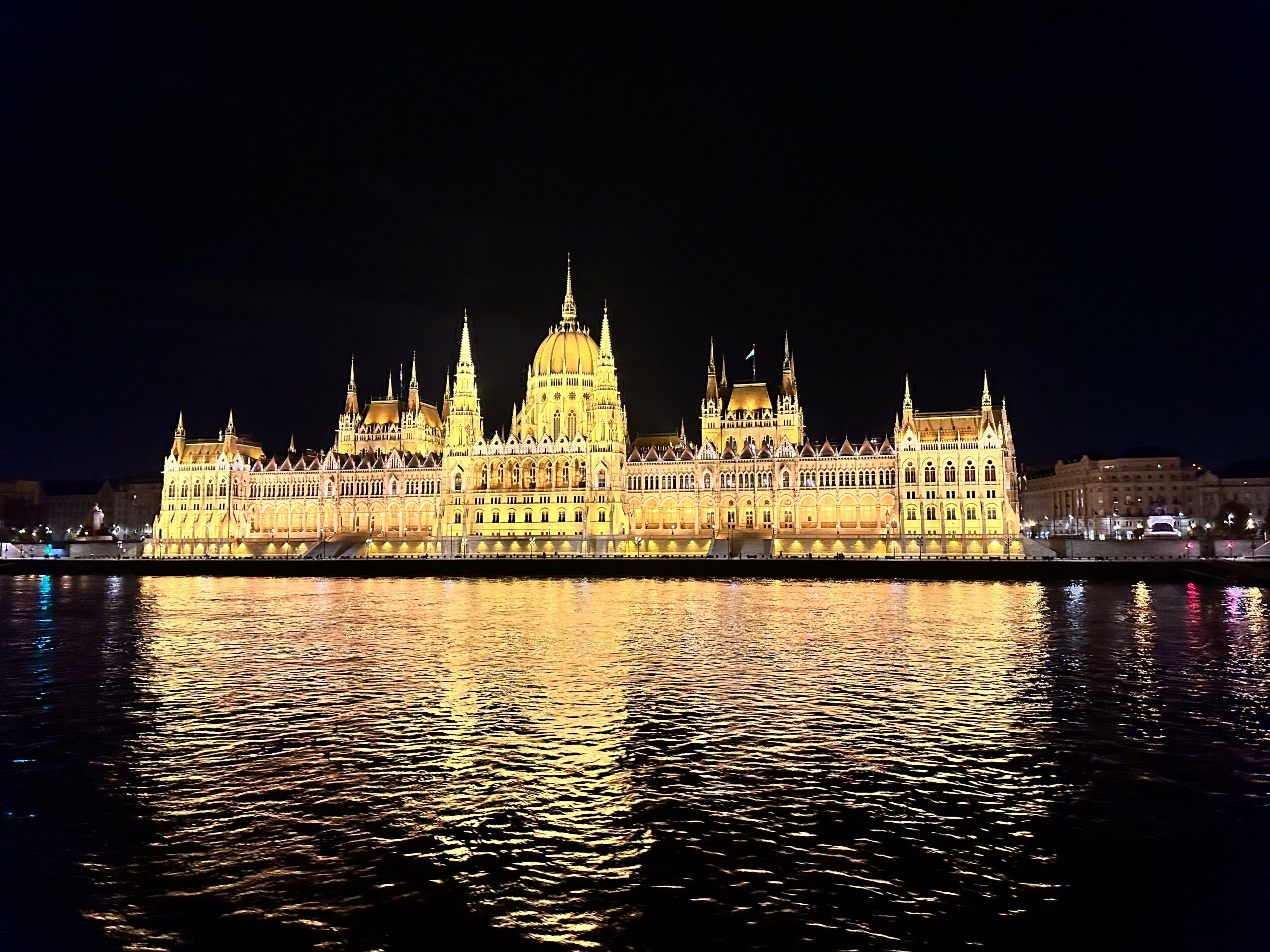 Night view of a illuminated historic building with multiple spires, reflected in water, against a dark sky.