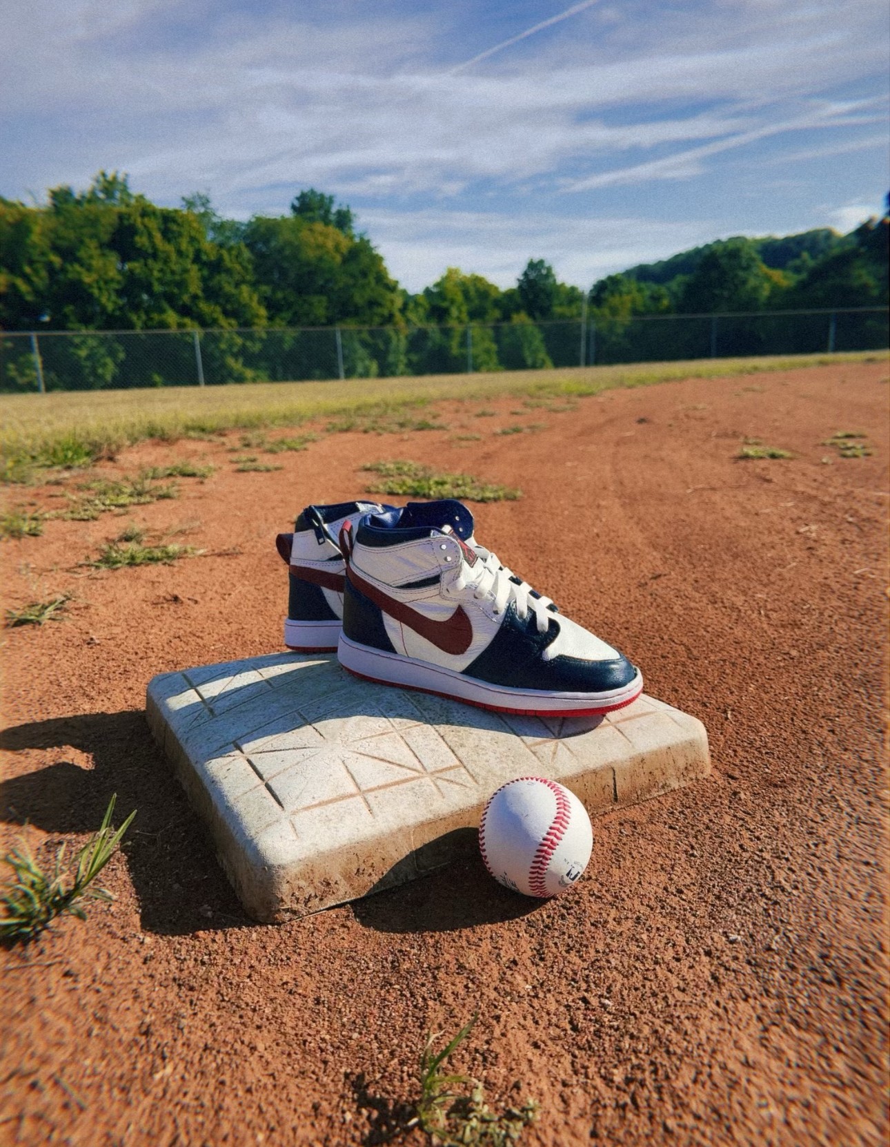 A baseball shoe on a raised platform with a baseball on the ground nearby, on a baseball field with trees and sky in background.