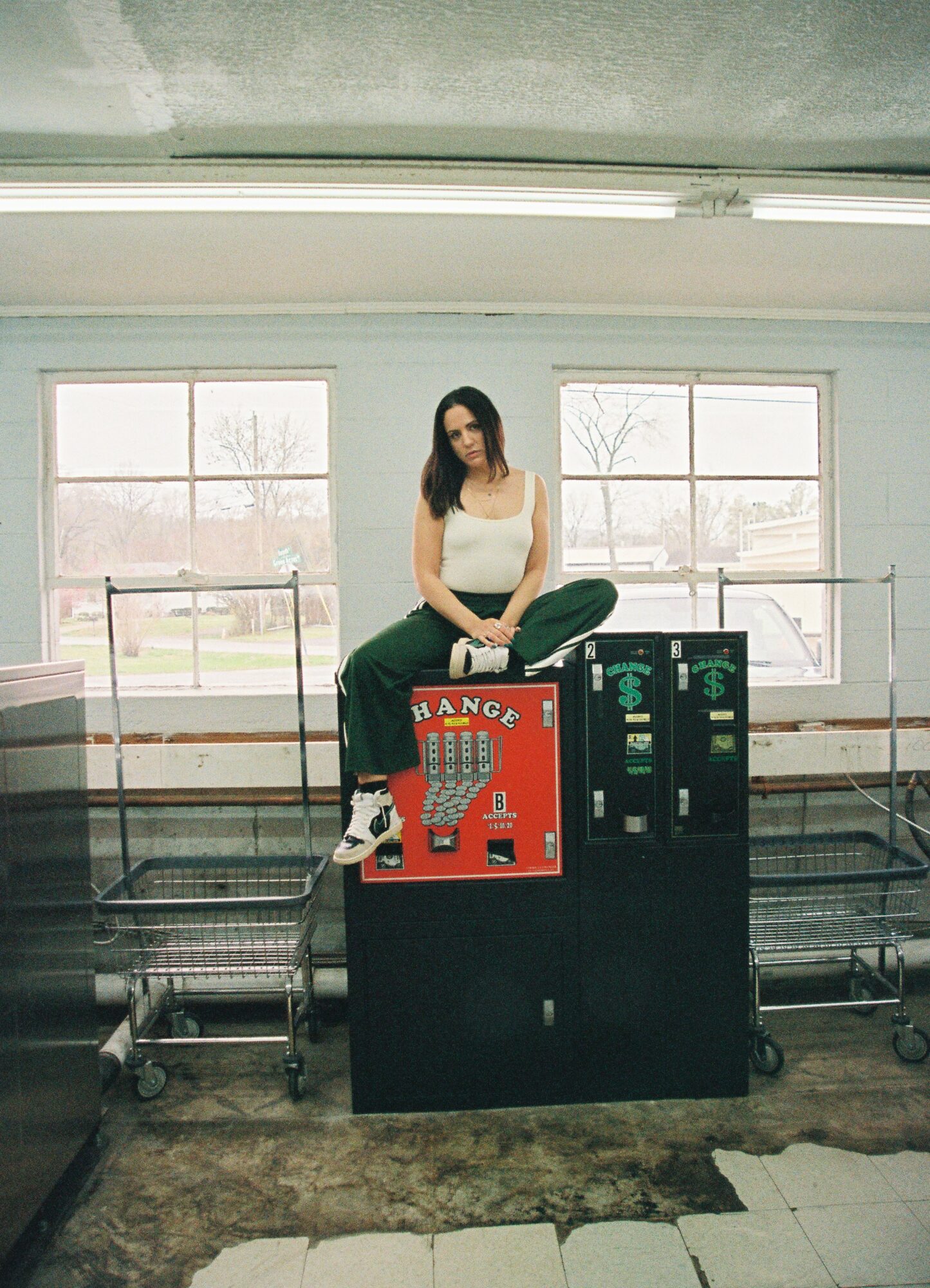 Woman sitting on a vending machine in a room with large windows and natural light, concrete floor, and metal carts nearby.