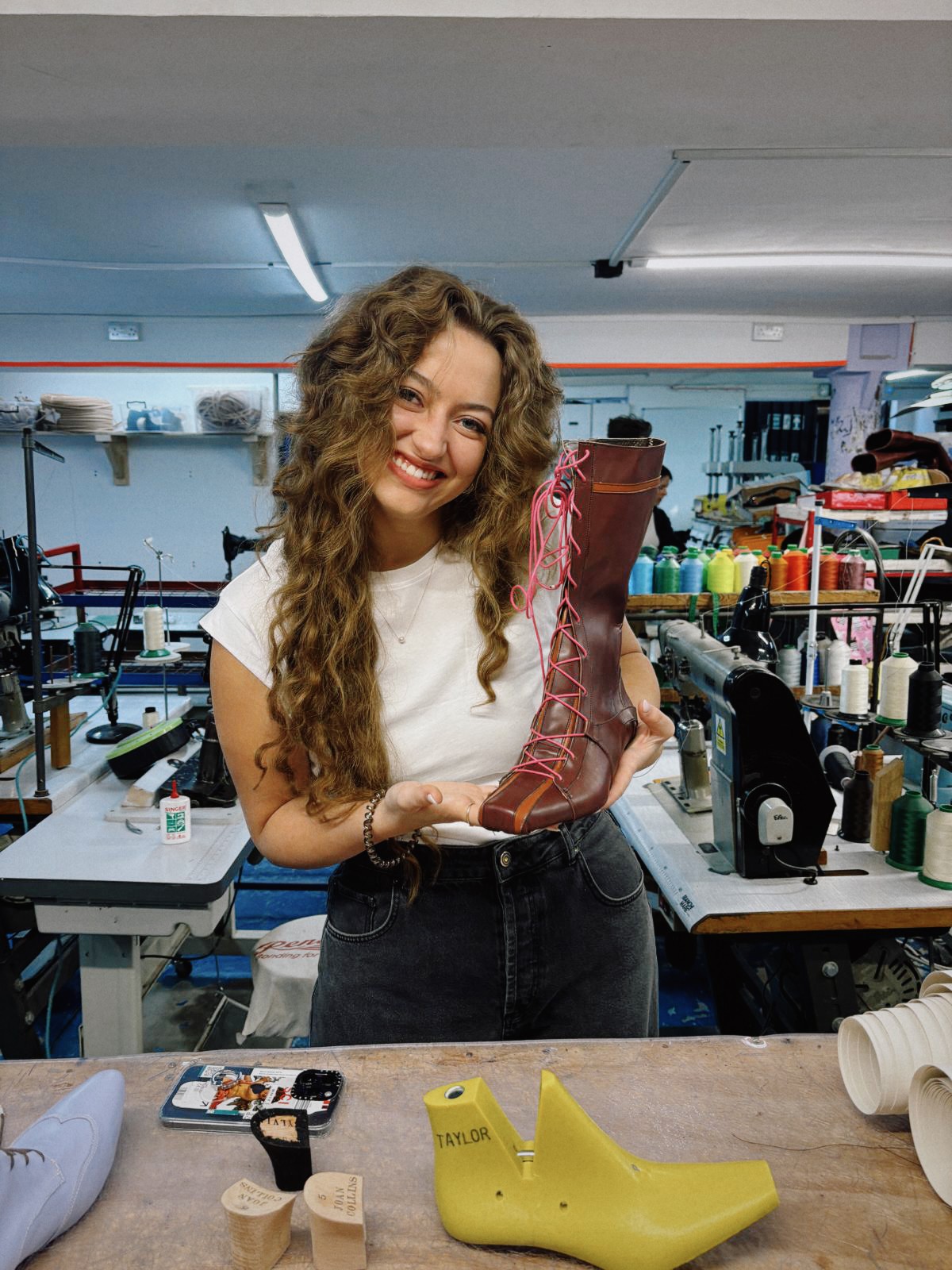 Young woman with long curly hair smiling, holding a red boot, in a workshop or store with sewing supplies.