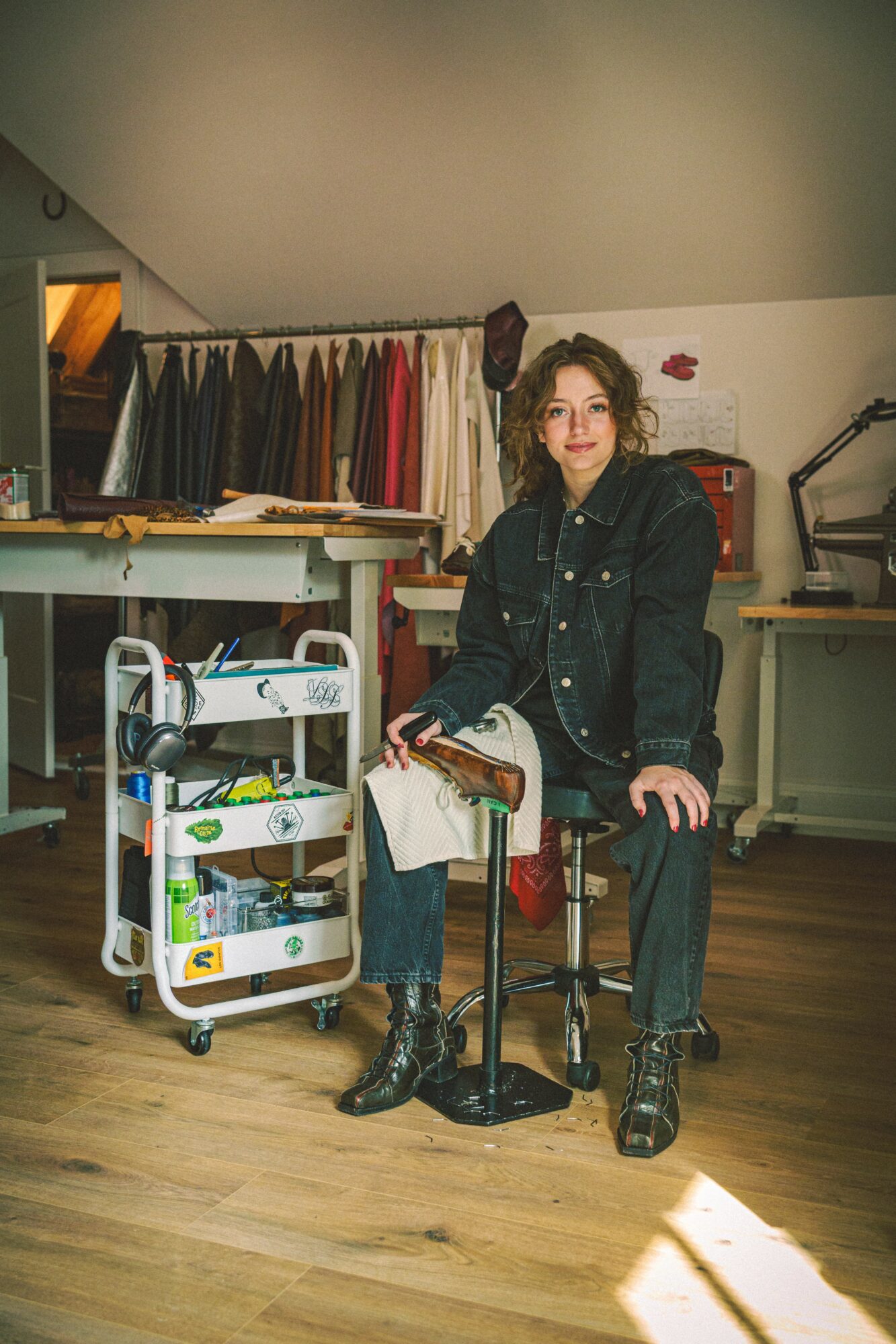 Woman sitting on a stool in a clothing store, surrounded by racks of colorful clothes and sewing equipment.