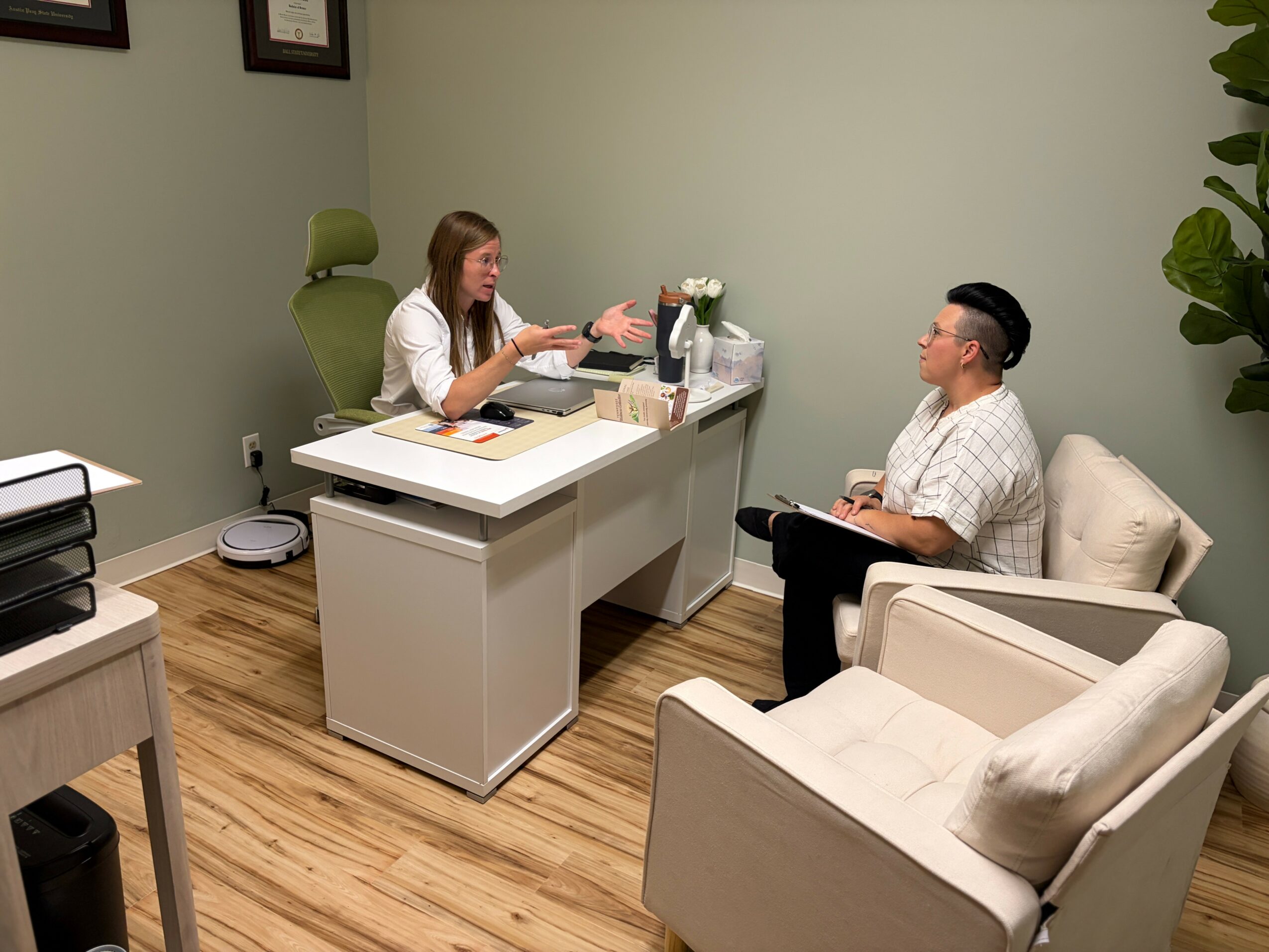 Woman and man sitting in an office, woman at desk talking, man on chair listening, office with green wall and wooden floor.