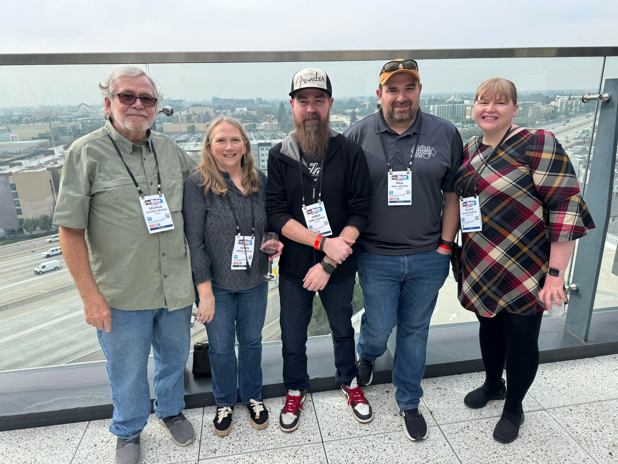 Five people standing on a balcony with a city view, smiling, wearing name tags, and casual clothing.