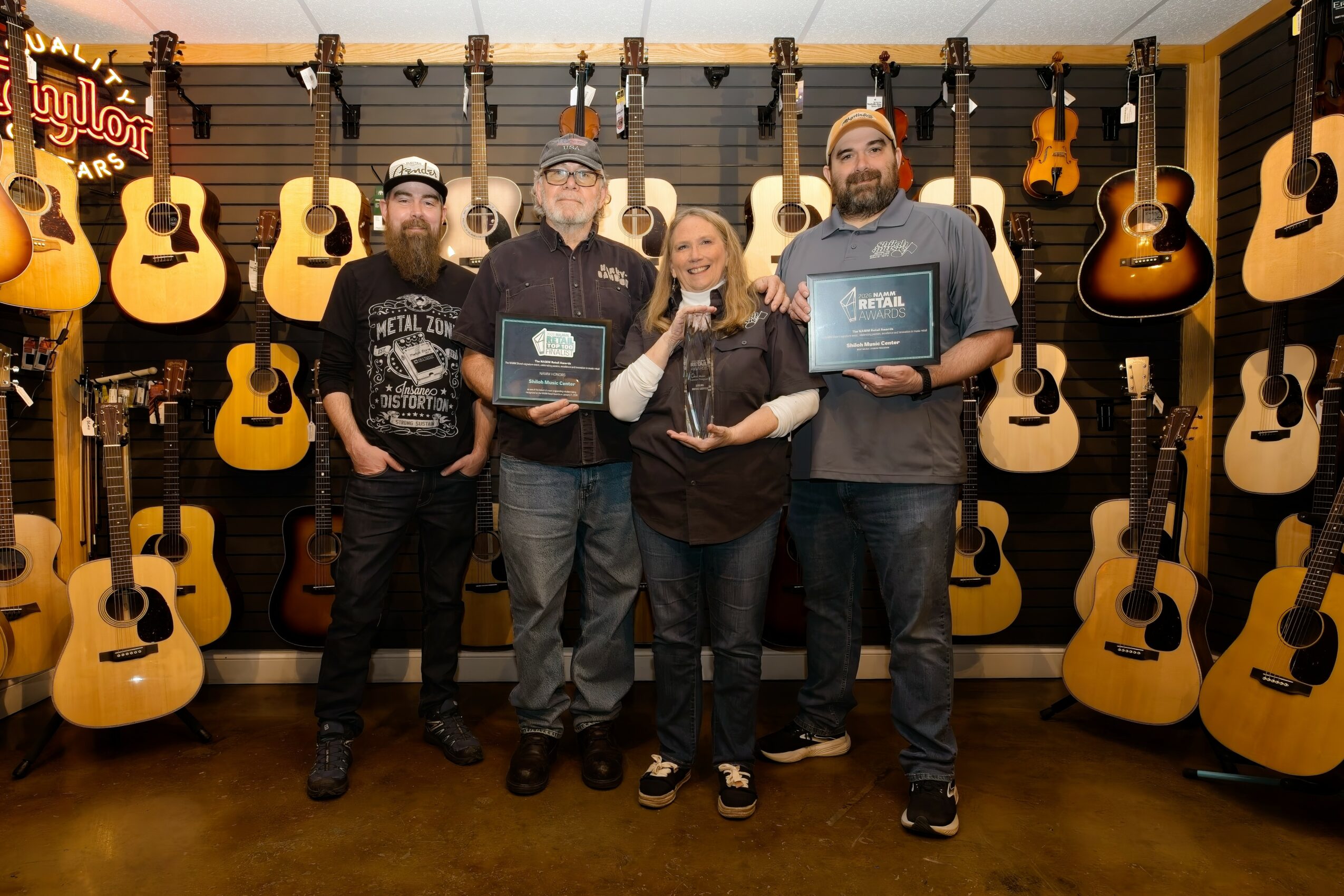 Four people standing in a guitar store, holding awards, surrounded by guitars on the wall behind them.
