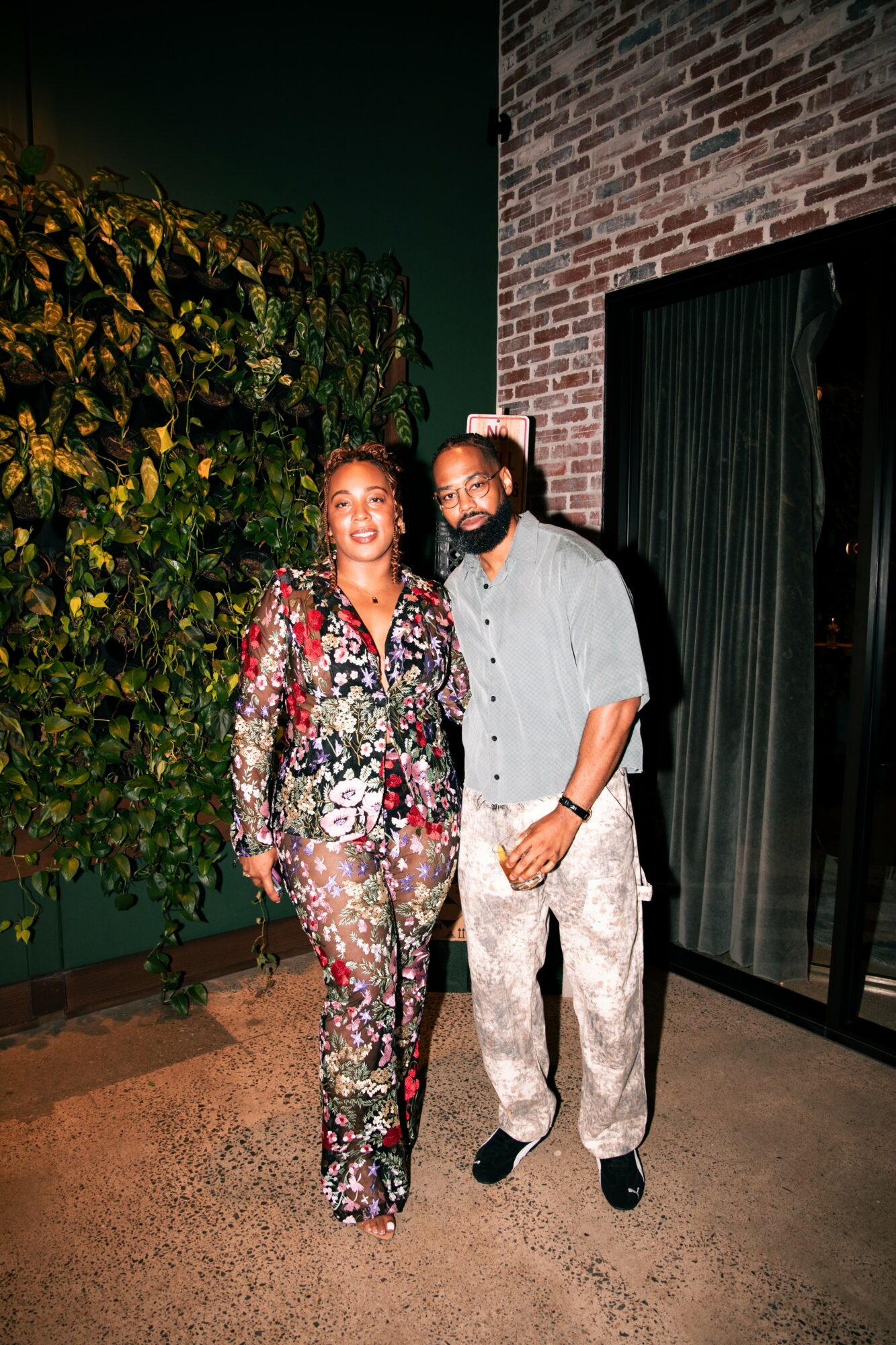 Two people standing outdoors near a brick wall and greenery, smiling at the camera.