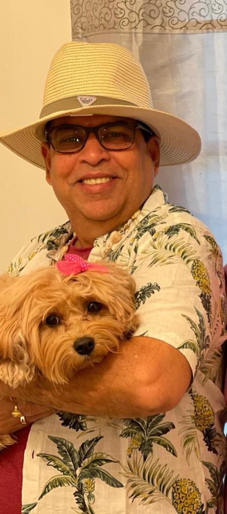 Person wearing a wide-brimmed hat and sunglasses holding a small dog, smiling indoors with curtains in the background.