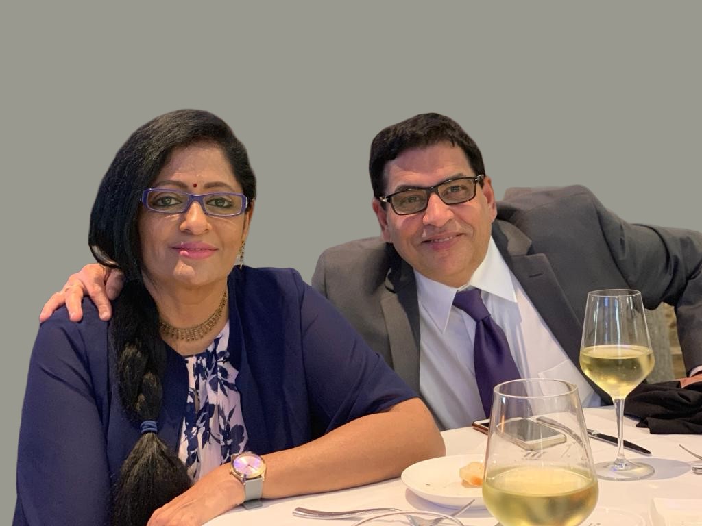 Two people sitting at a table with wine glasses, smiling, against a plain background.