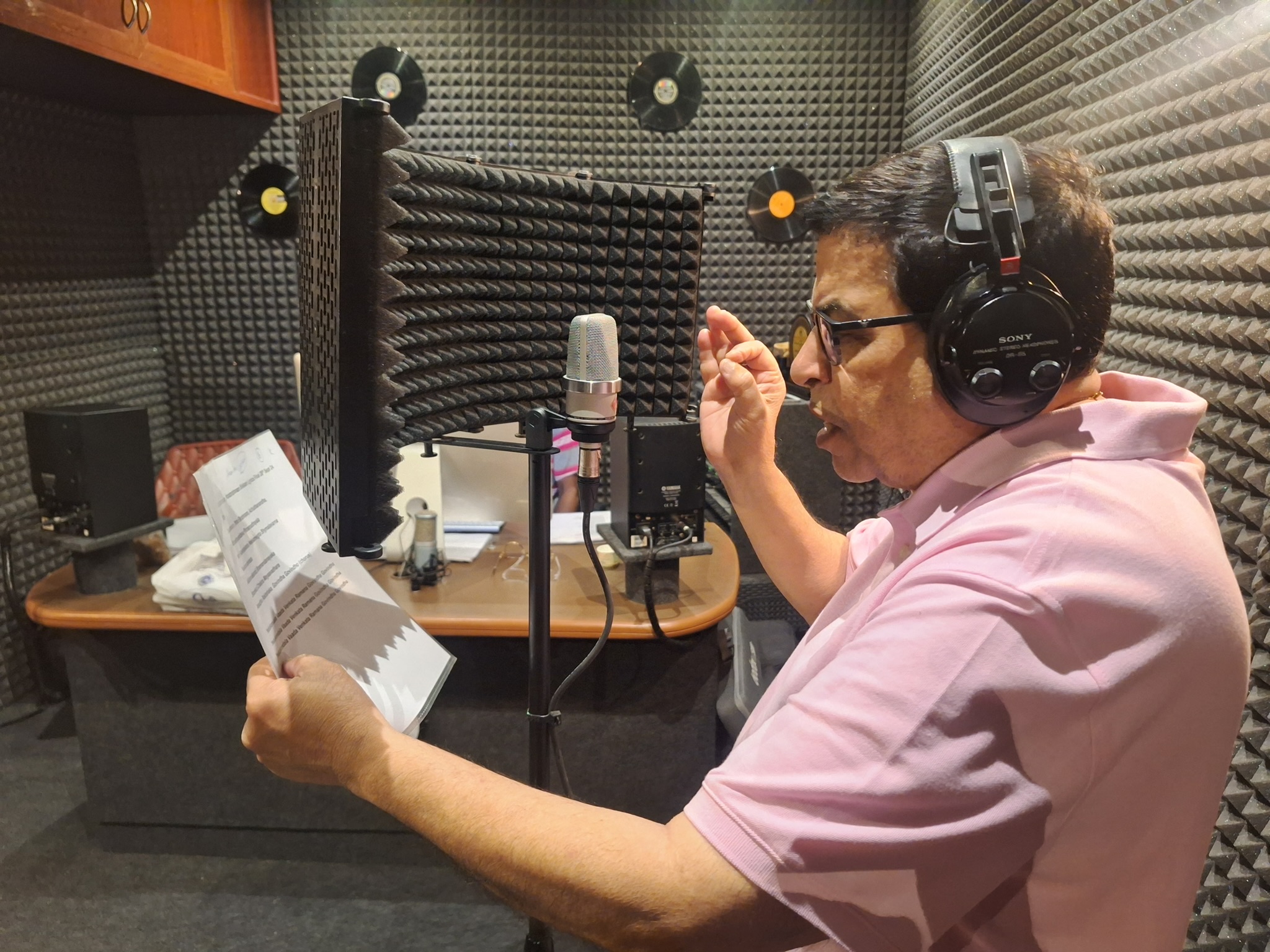 Man in pink shirt with headphones reading script in recording studio with soundproofing panels.