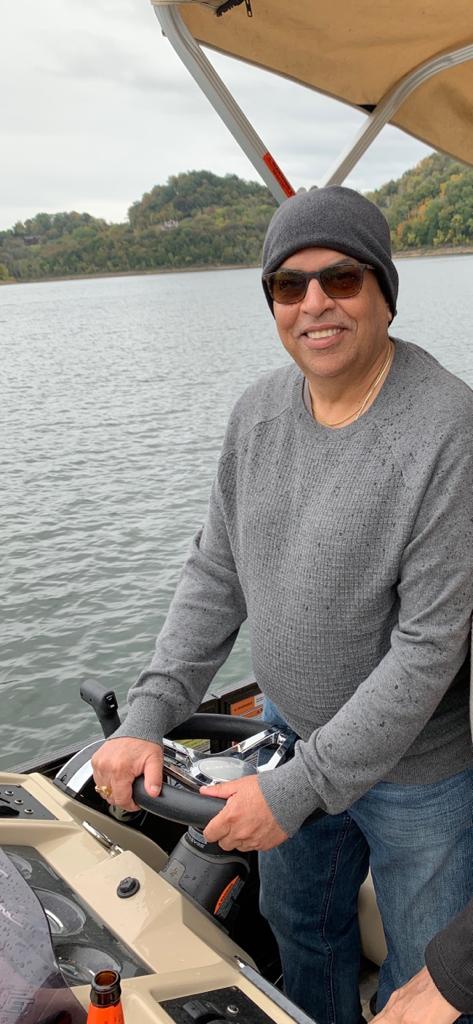Man wearing sunglasses and a gray sweater, smiling, standing on a boat with water and trees in background.