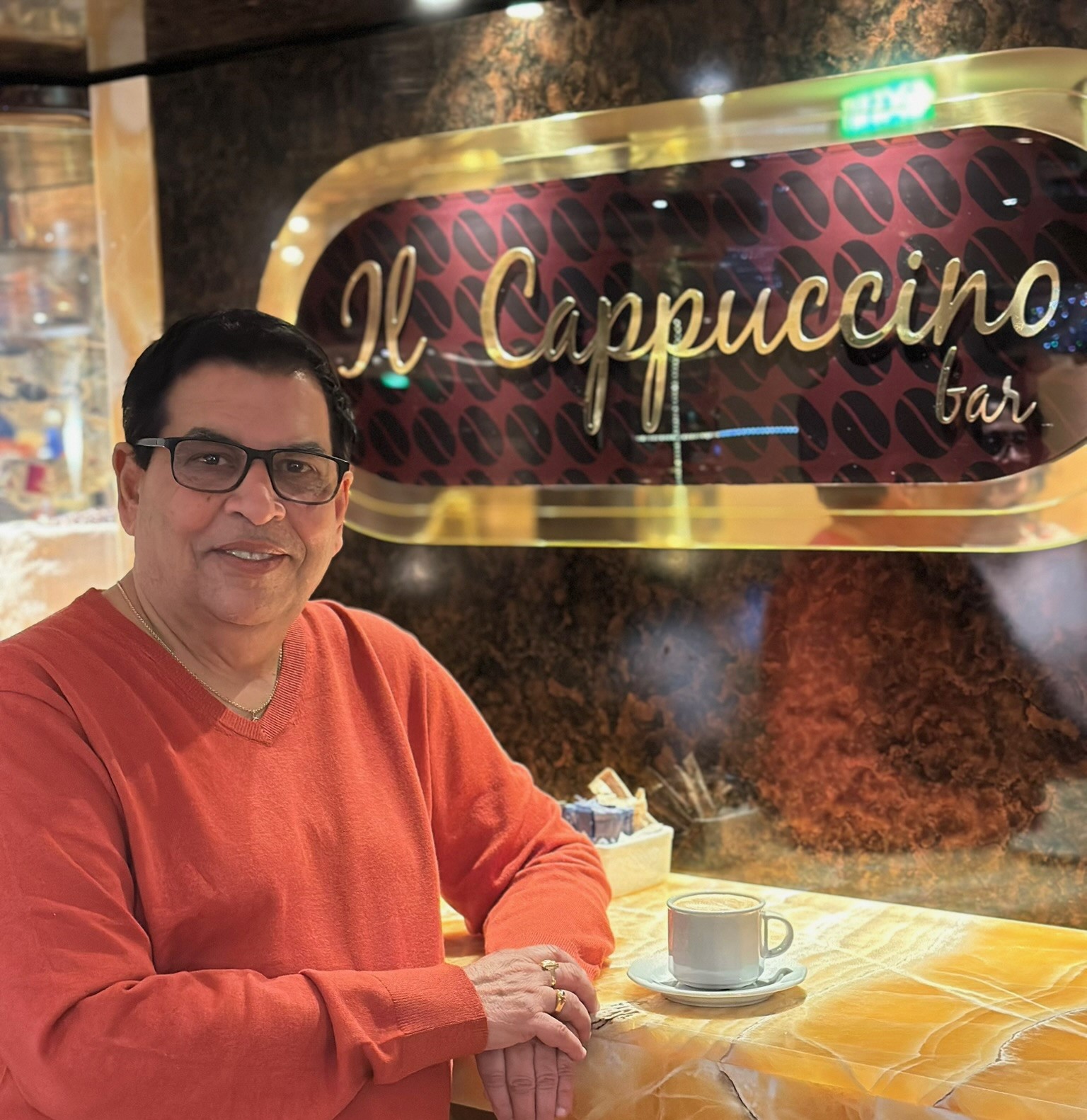 Man in red sweater sitting at a table with coffee cup in front, behind a sign that reads 'Il Cappuccino bar'.