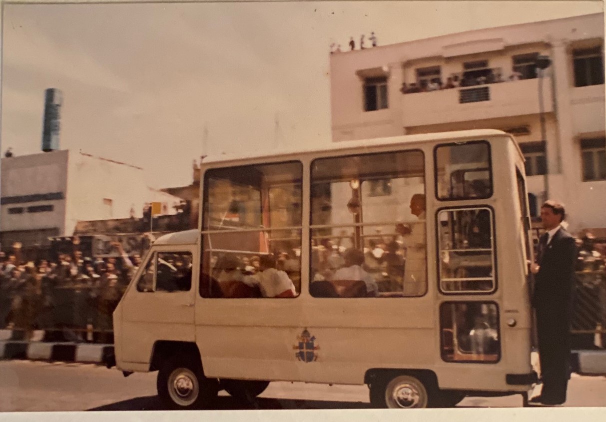 A small bus with large windows parked in front of a building, with a person standing nearby, in a city scene.