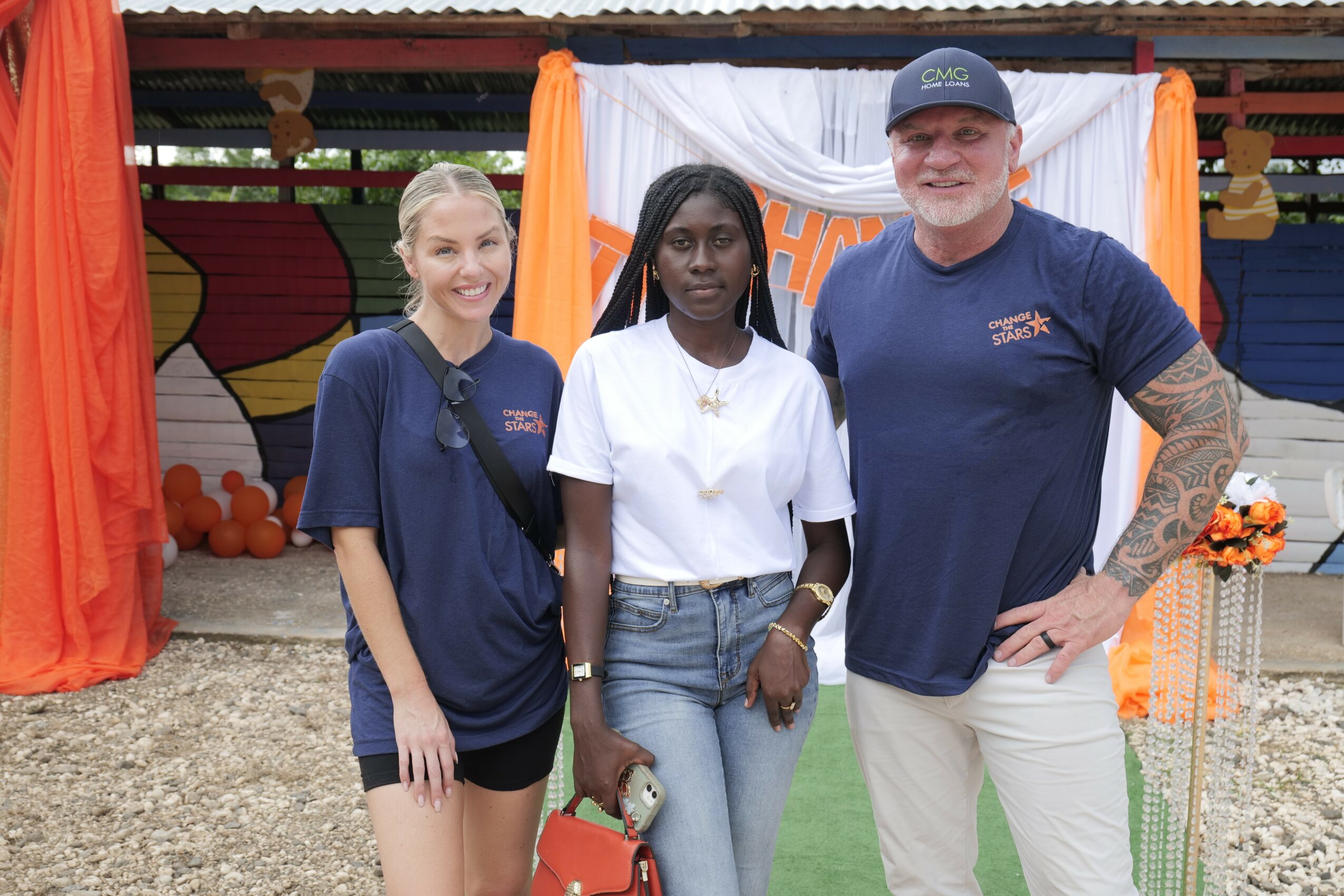 Three people standing outdoors in front of colorful decorations, smiling, with a gravel ground and a red building in the background.