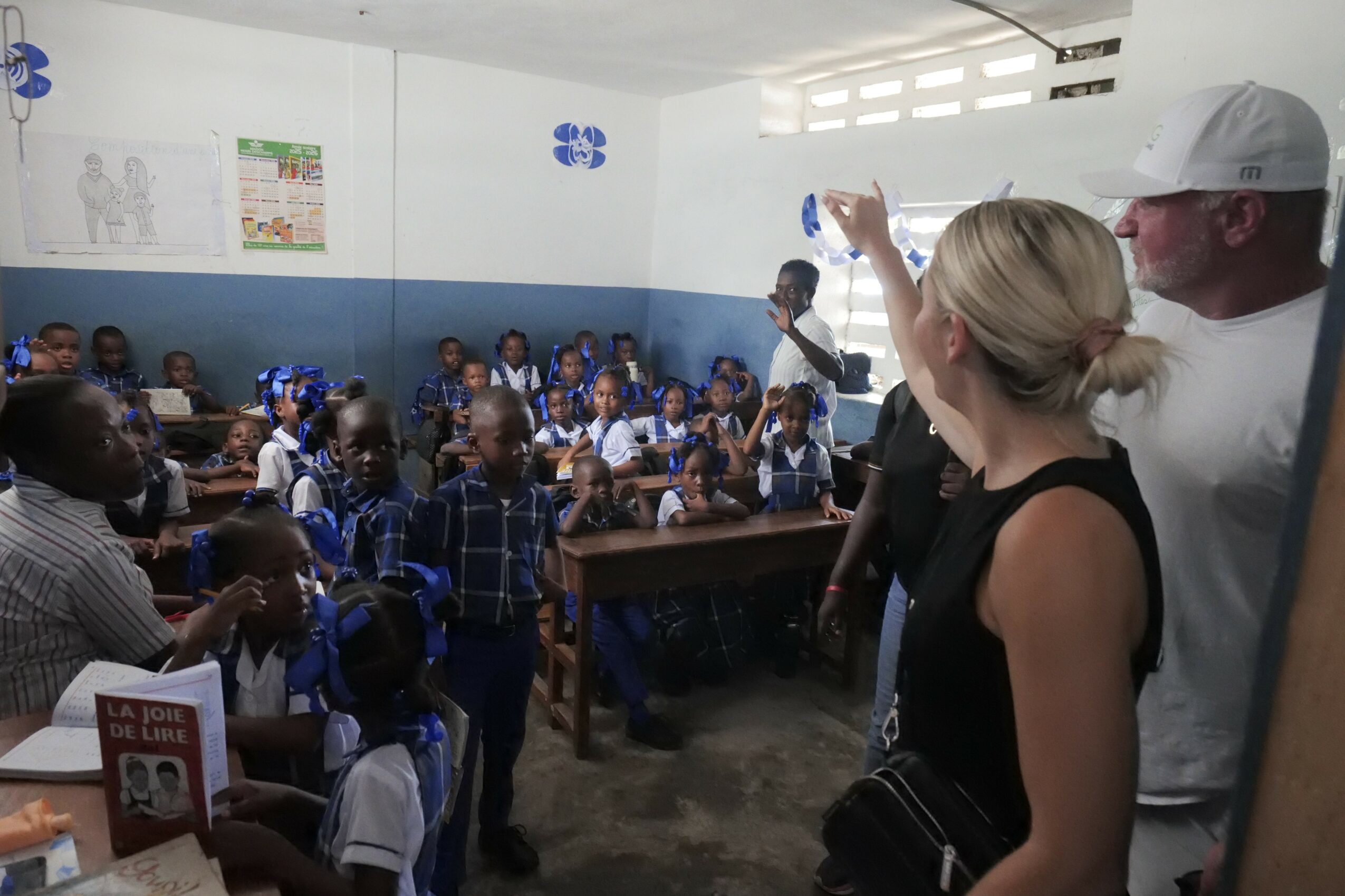 Group of children in school uniforms sitting at desks in classroom with two adults standing nearby, one woman raising her hand.