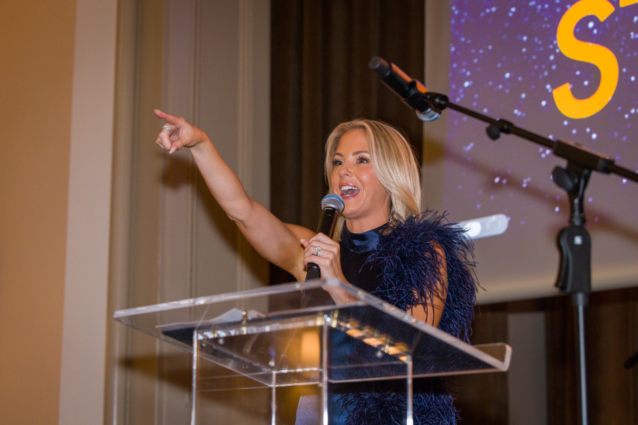 Woman speaking at a podium, pointing with her right hand, holding a microphone, with a starry background.