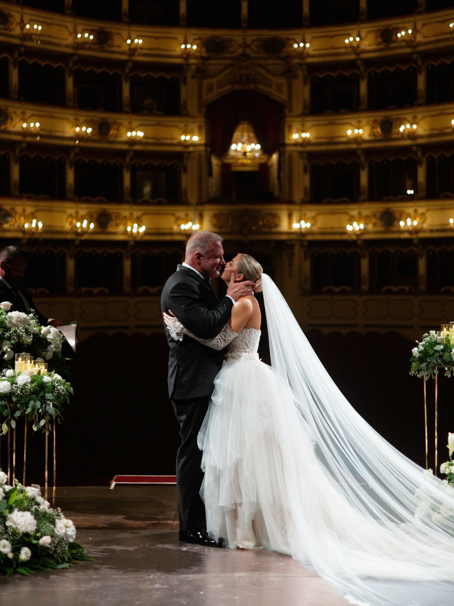 A bride and groom embrace on stage in a grand theater with multiple balconies and ornate architecture, decorated with flowers.