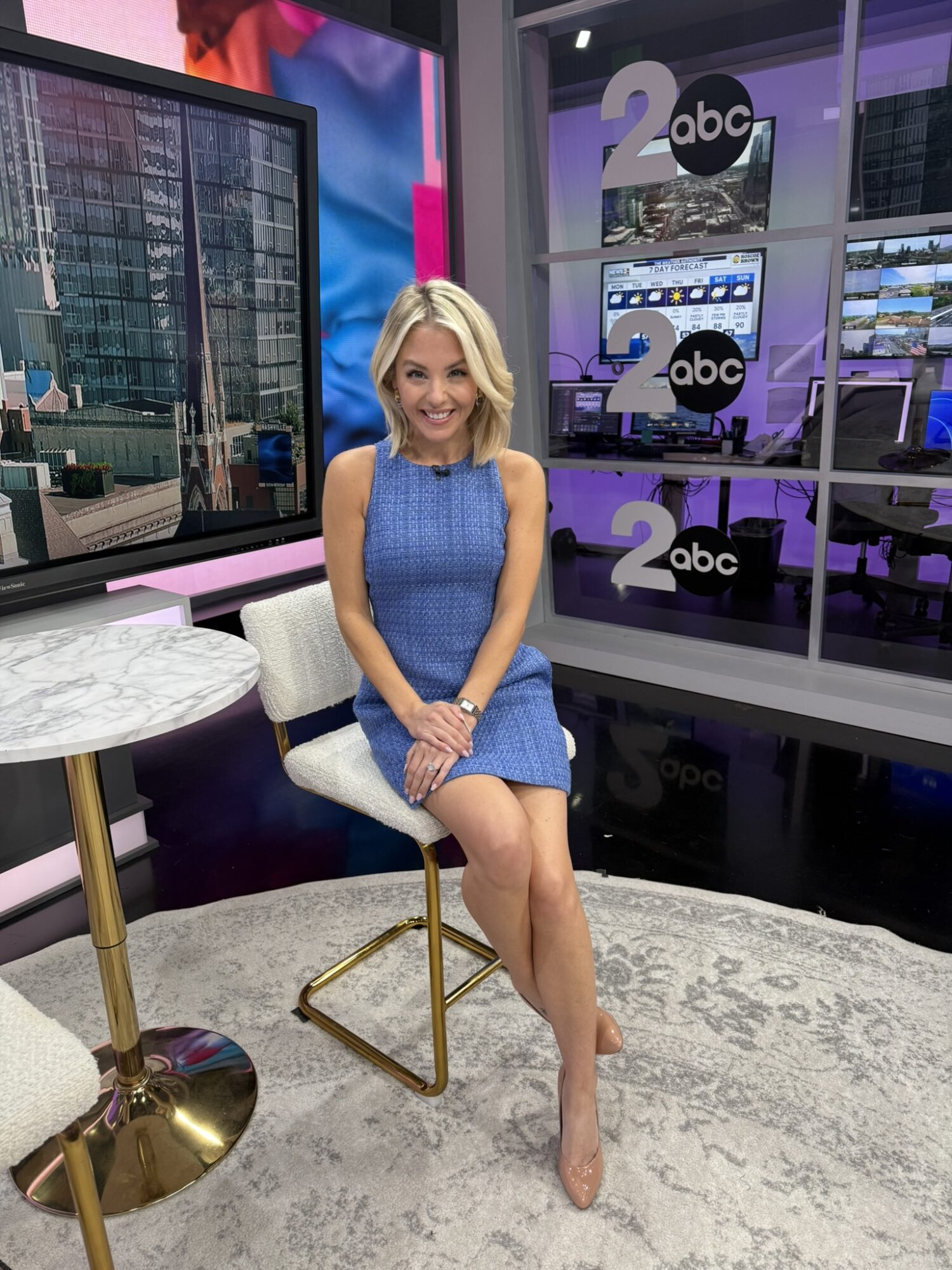 Woman with blonde hair in a blue dress sitting on a white chair in a TV studio, with screens and windows behind her.