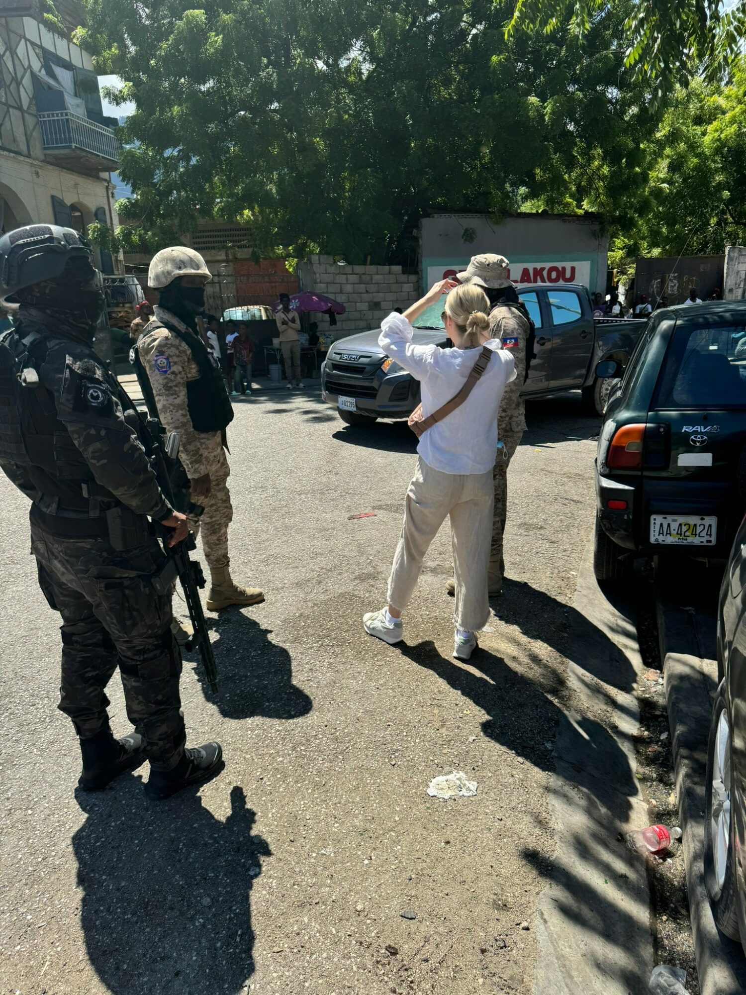 Group of people, including a woman with a backpack, standing on a street with parked cars and trees in background.