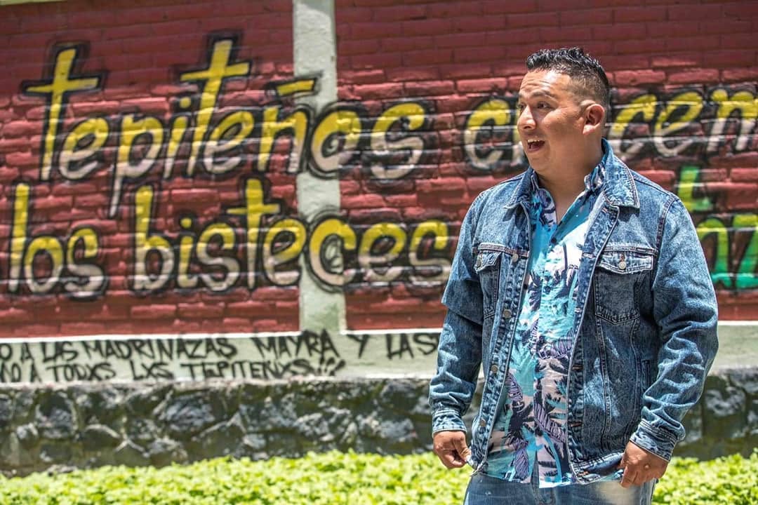 Young man standing outdoors in front of a colorful graffiti wall, wearing a denim jacket and patterned shirt.