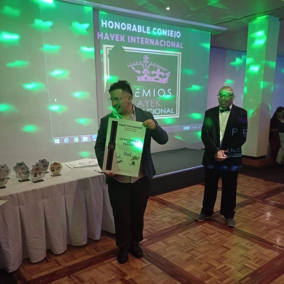 Person holding a framed award on stage at an event with a large screen behind displaying text and logo, green lighting effects.