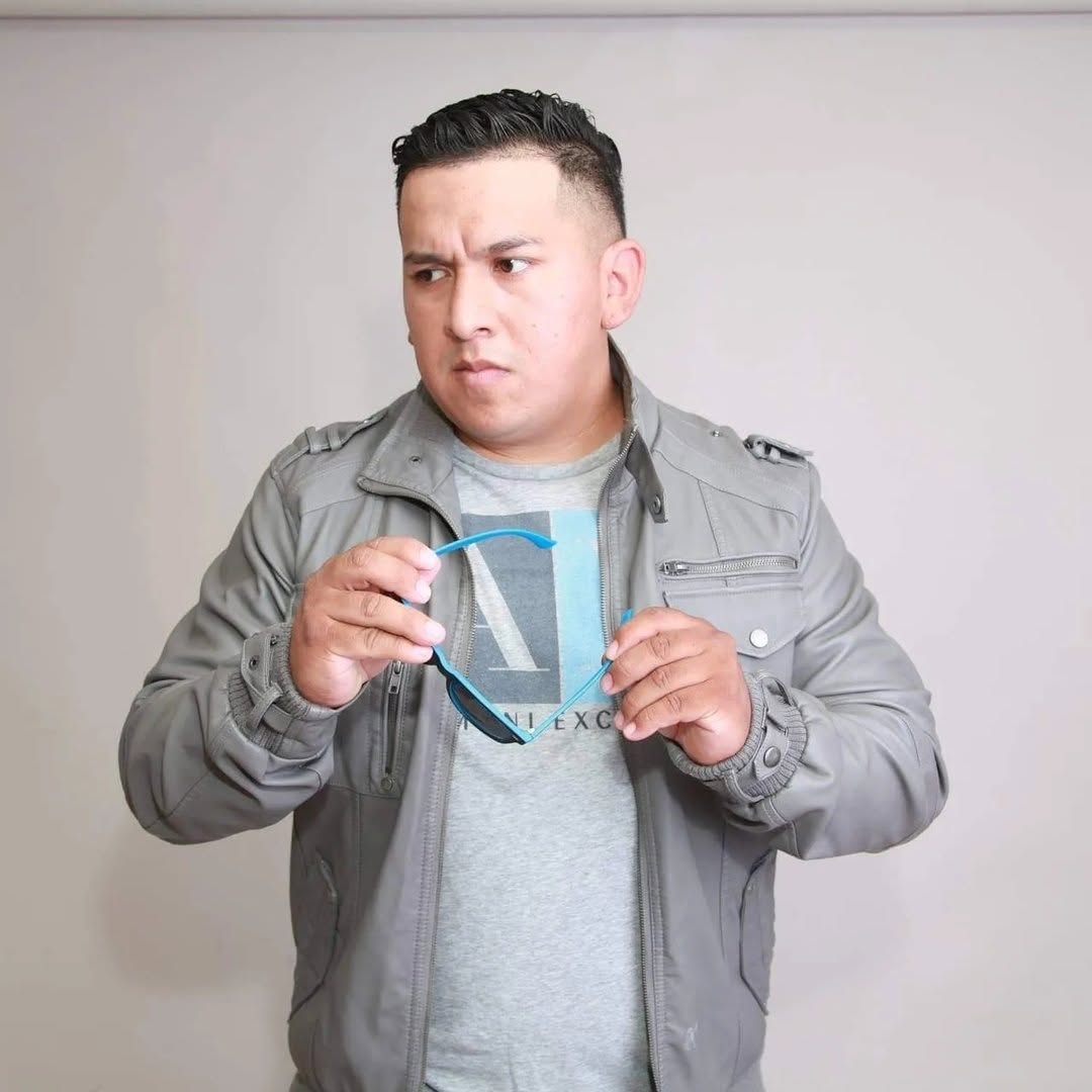 Man holding glasses, looking to the side, wearing a gray jacket and t-shirt, against a plain background.