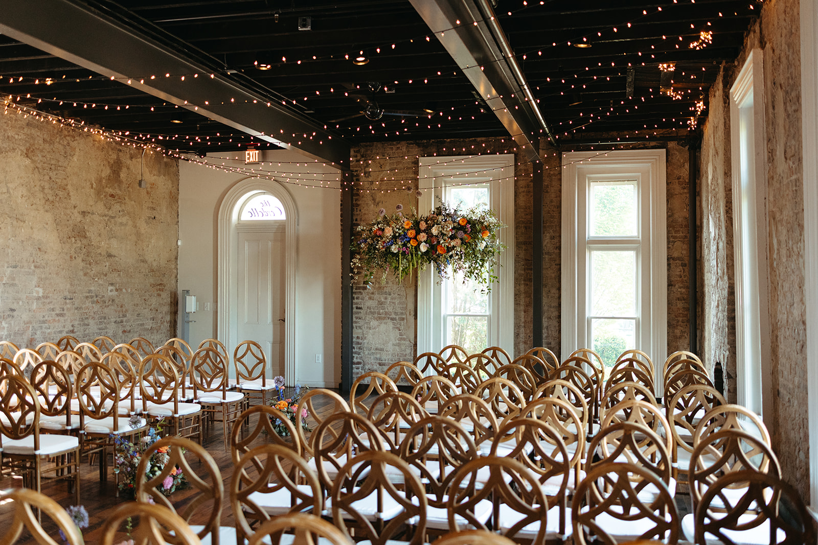 Room with wooden chairs arranged in rows, decorated with string lights and floral arrangements, large windows, and a door.