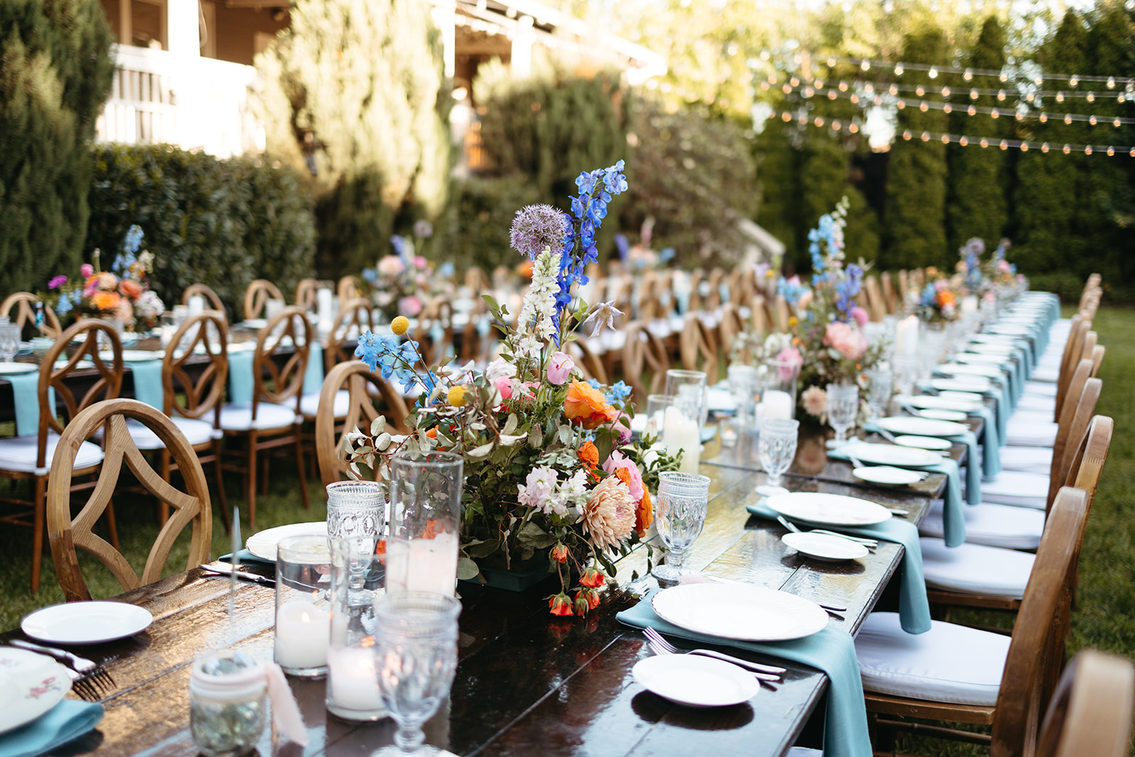 Long outdoor table set with plates, glasses, and floral centerpieces, surrounded by chairs, in a garden setting with trees and string lights