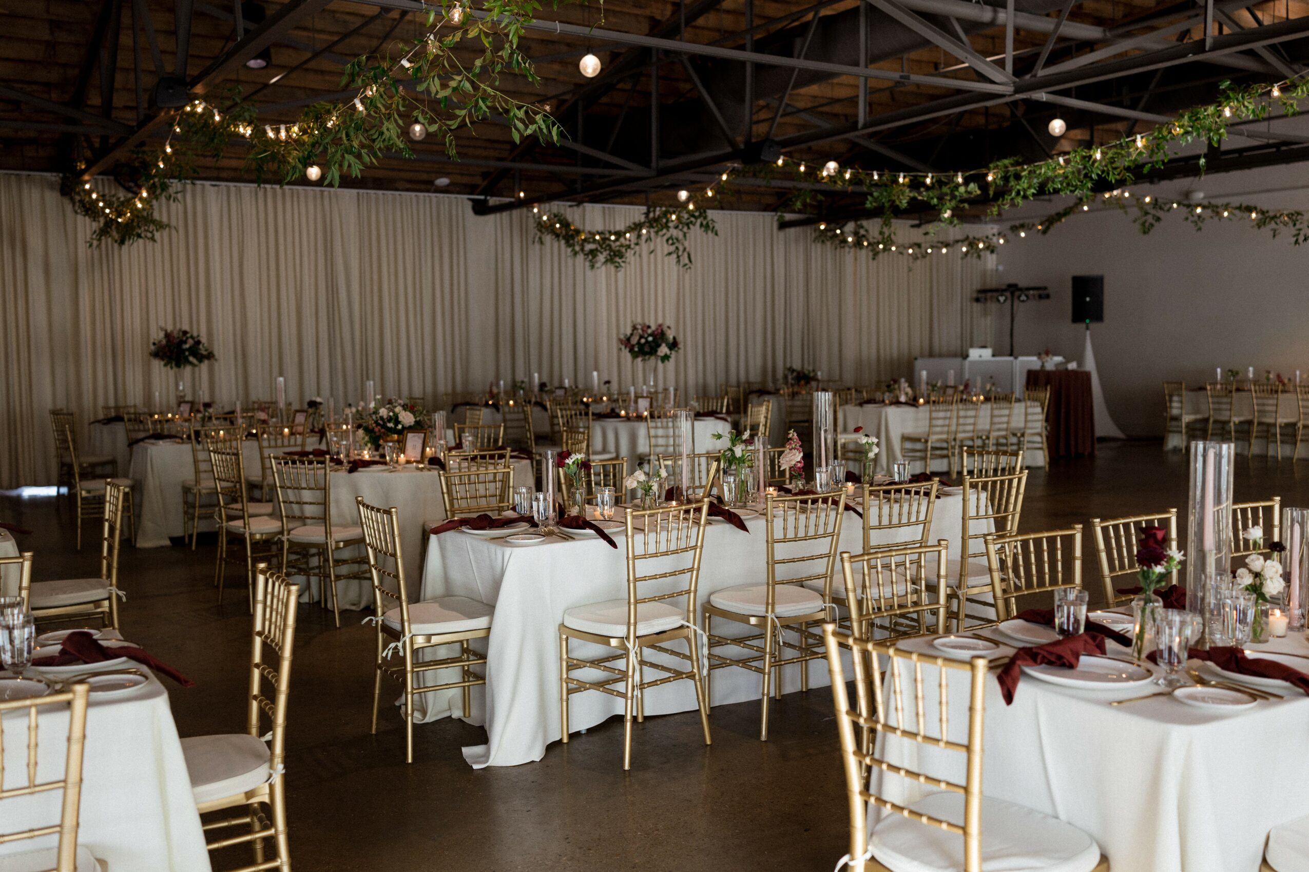 Wedding reception setup with round tables, white tablecloths, gold chairs, and string lights overhead.