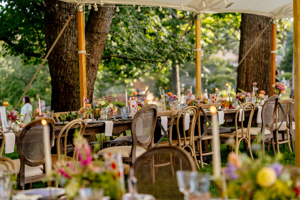 Long outdoor table set with chairs, flowers, and decorations under a canopy, surrounded by trees and greenery.