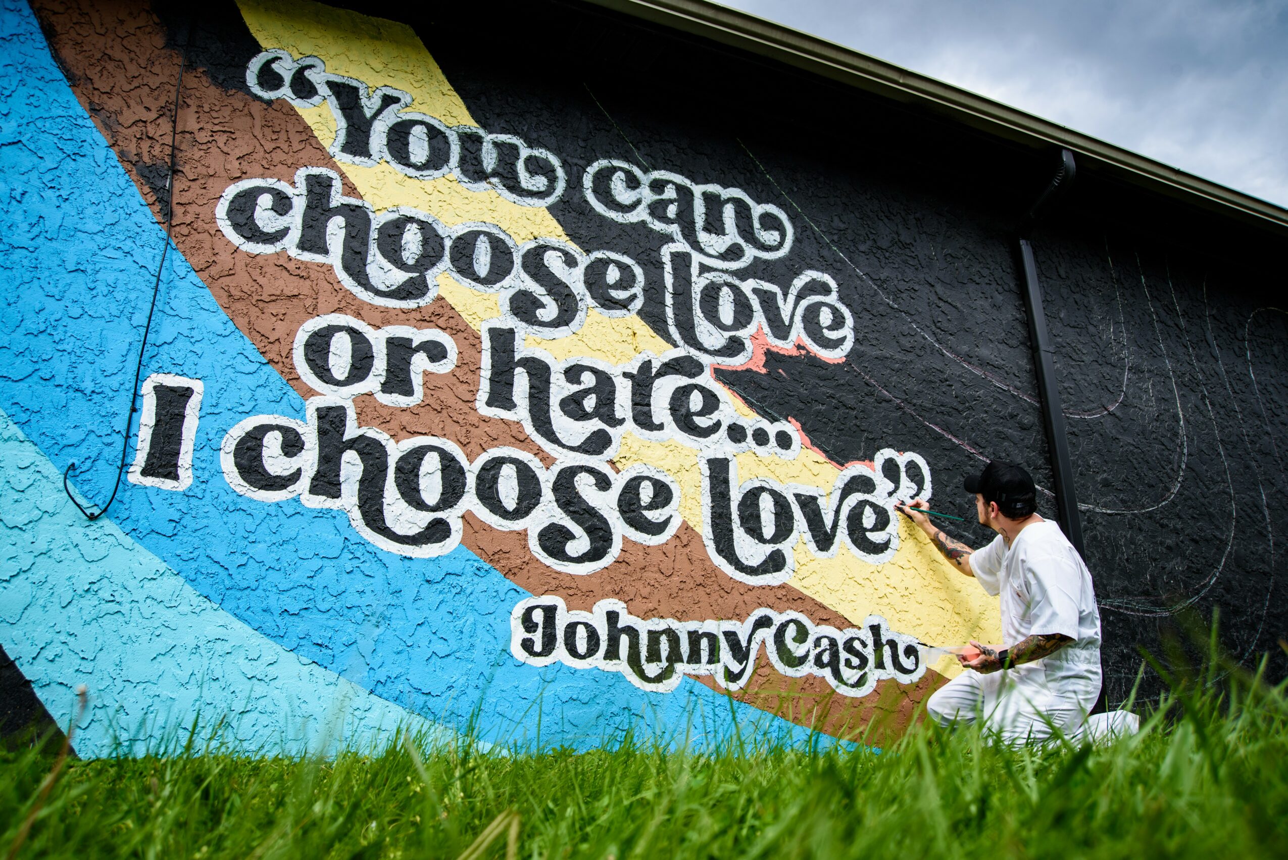 Person painting a mural with a quote about love and hate, on a black wall, with grass in foreground.