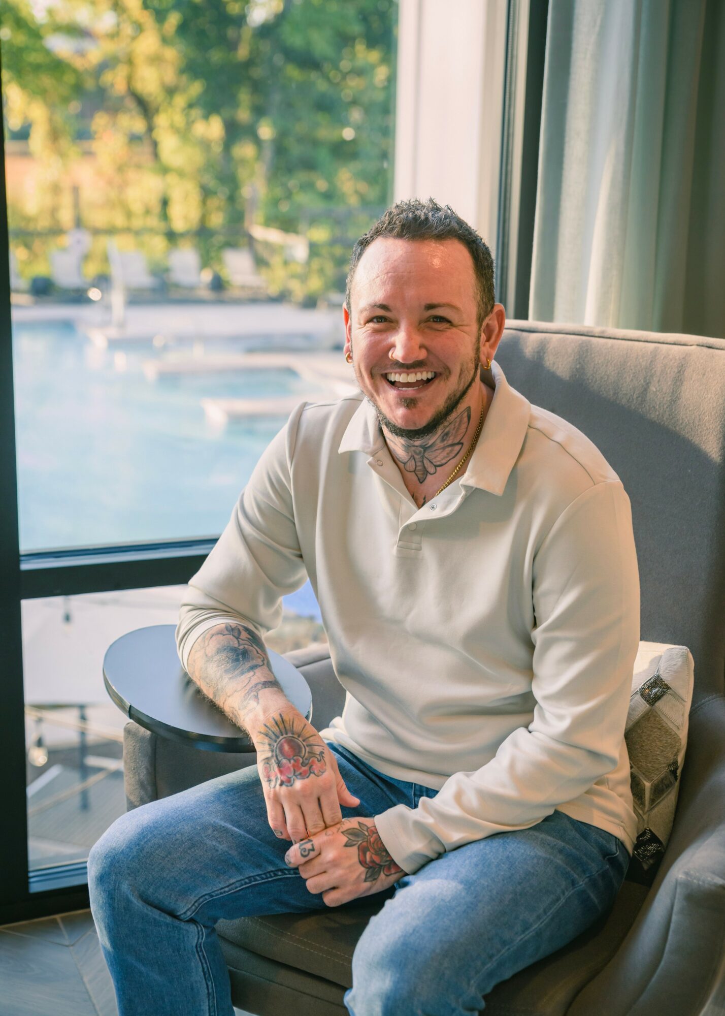 Smiling man with tattoos sitting by a window, wearing a white shirt and jeans, with trees outside.