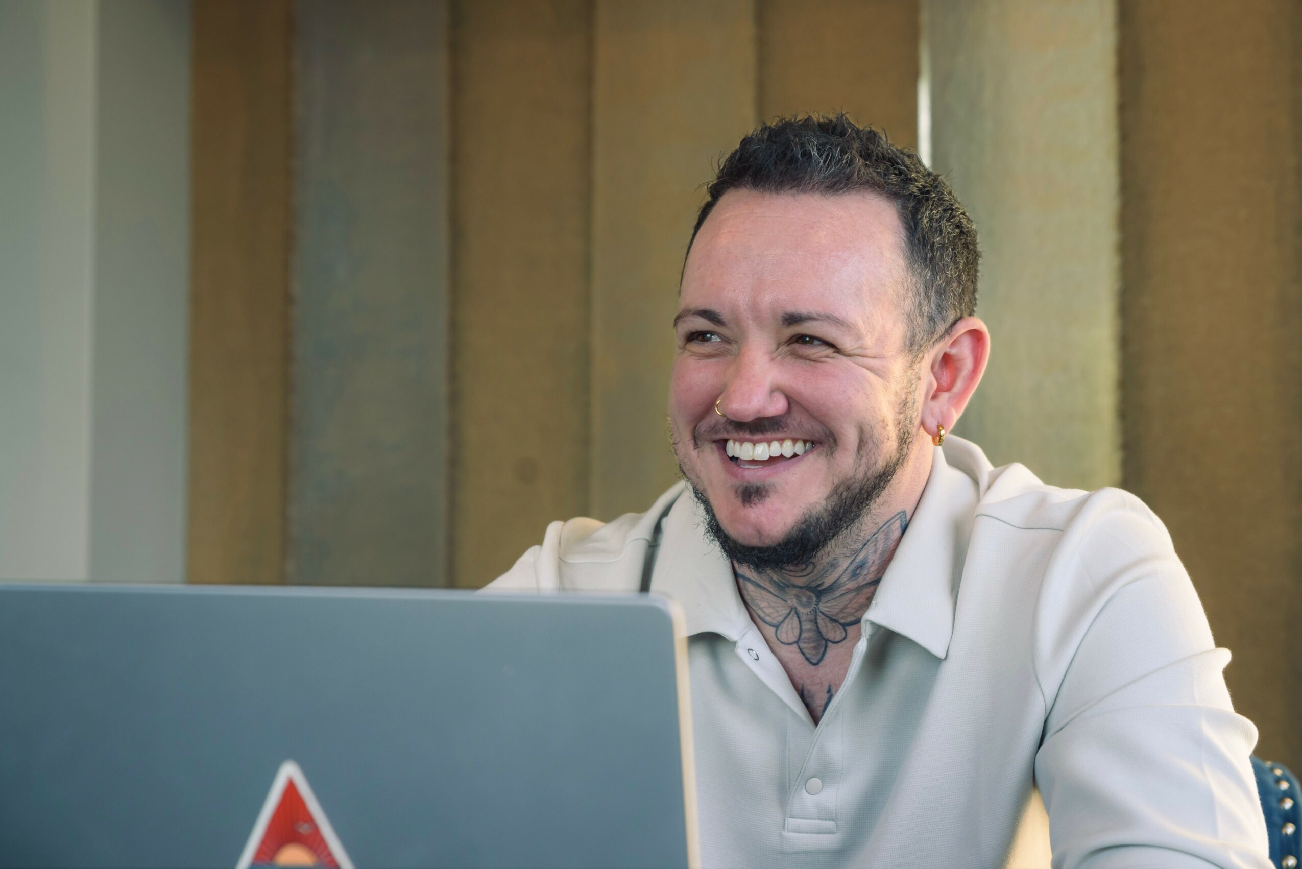 Smiling man with tattoos on neck, wearing a white shirt, sitting in front of a laptop, in a room with vertical blinds.
