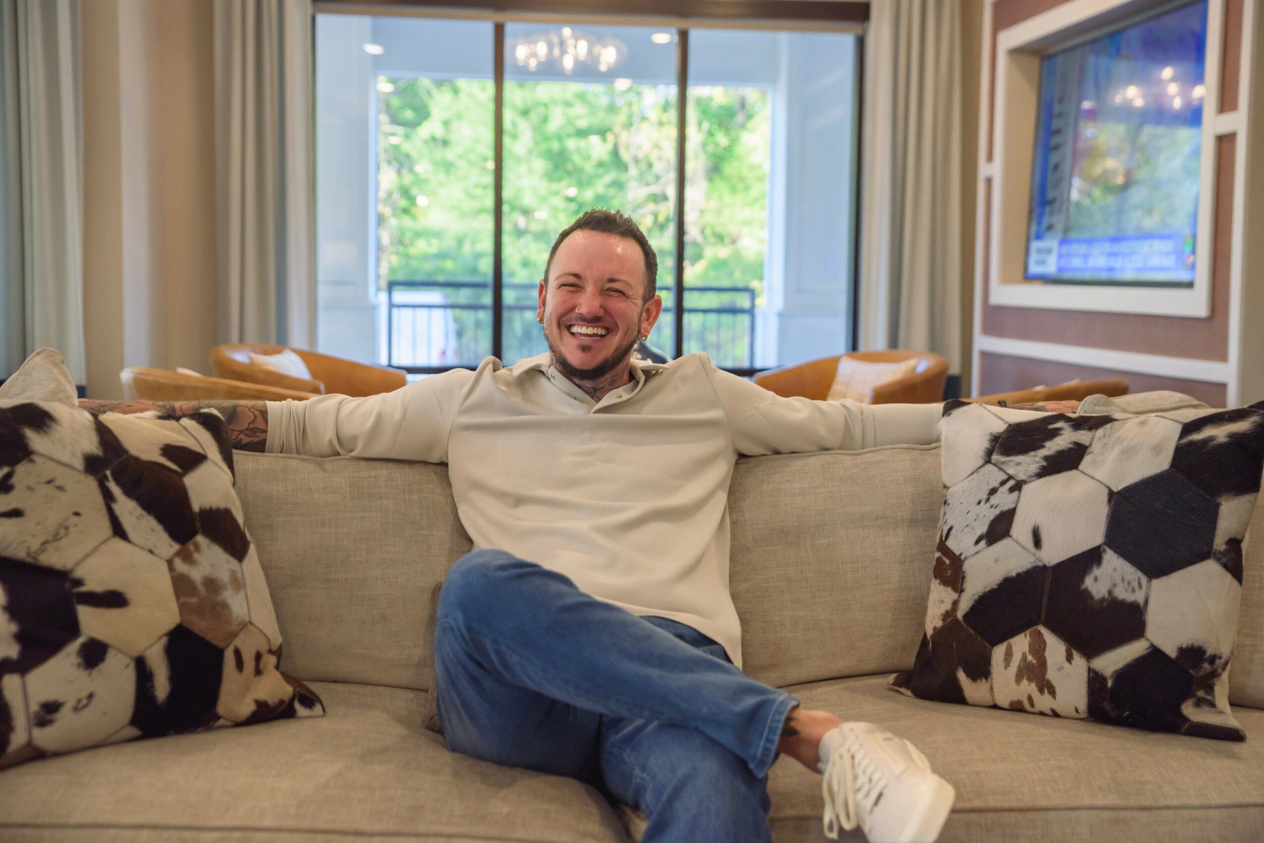 Man sitting on a sofa with arms outstretched, smiling, in a bright room with large windows and patterned pillows.
